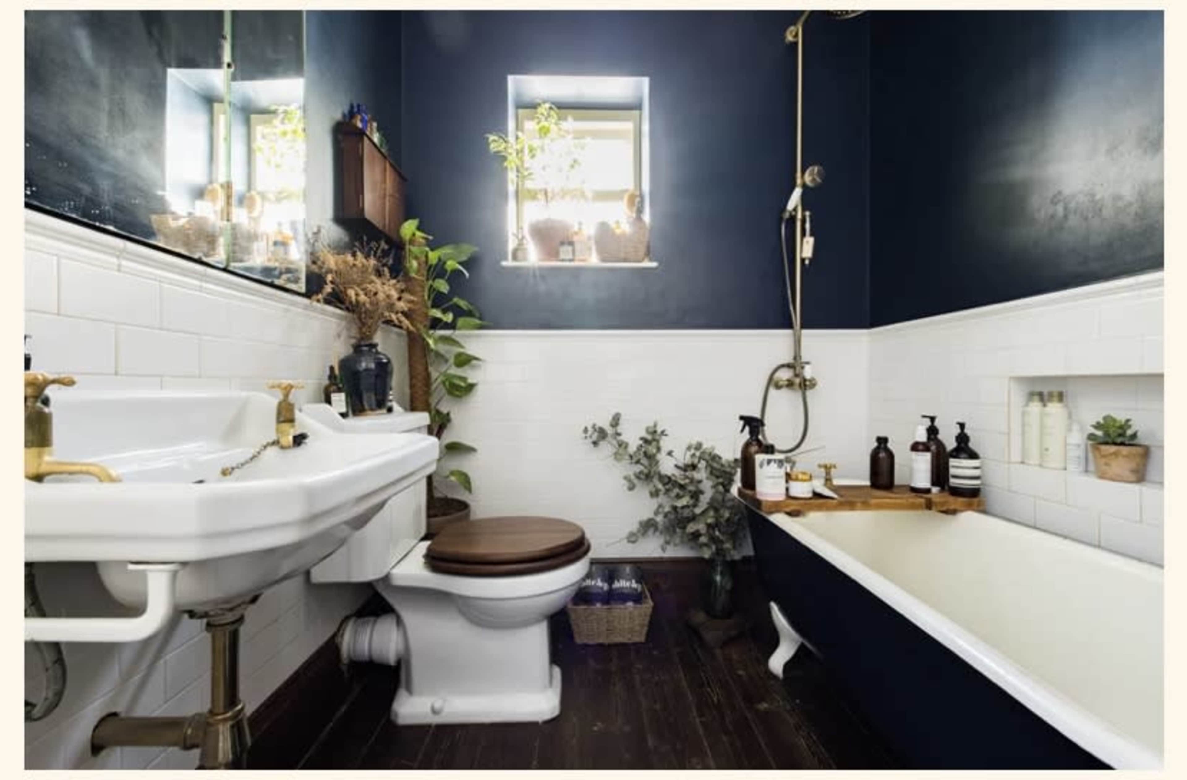 A compact bathroom featuring white tiled walls, a wooden floor, a porcelain toilet, a vintage-style sink, a clawfoot bathtub, and shelves with plants and toiletries.