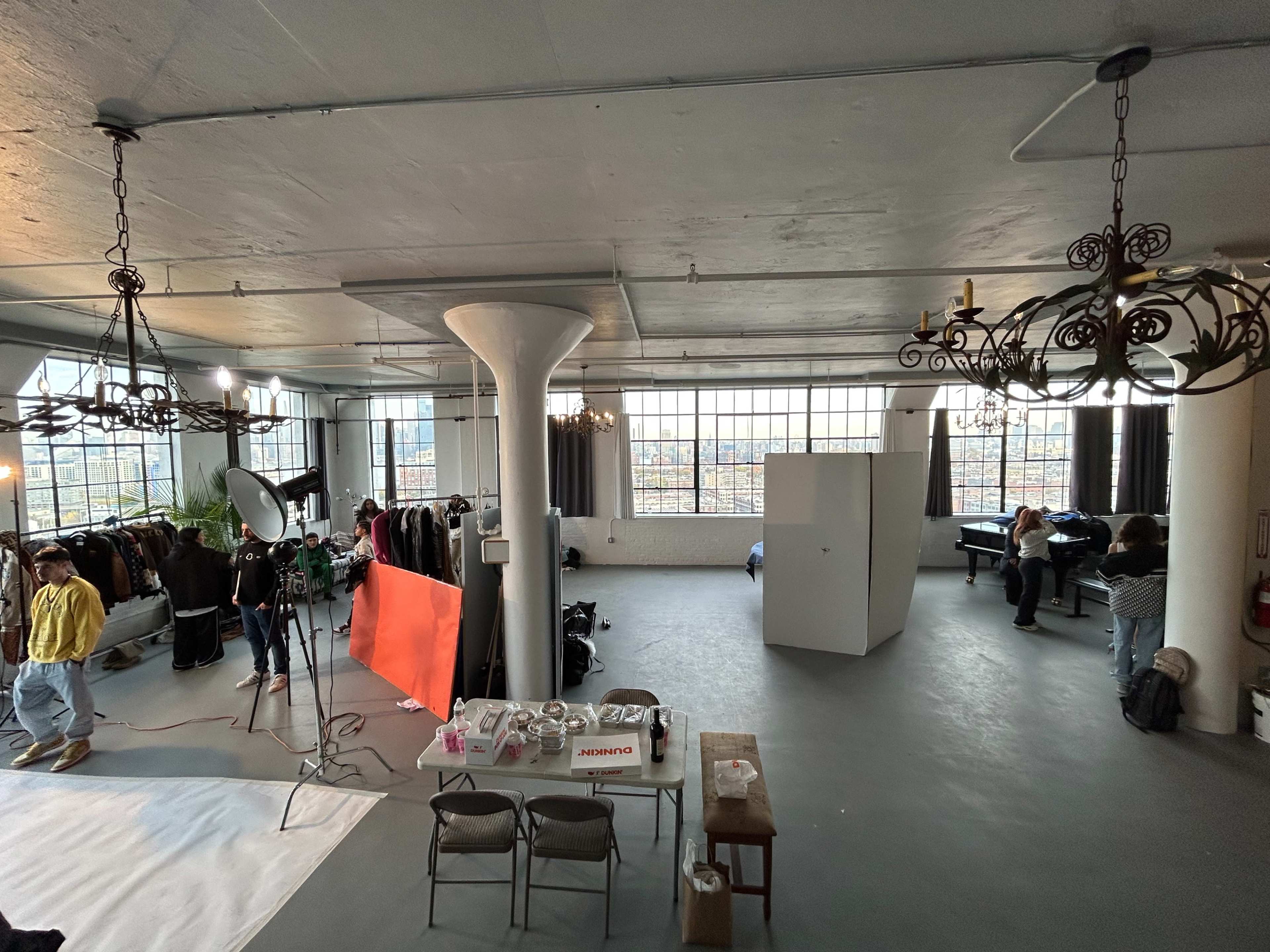 A spacious industrial studio with large windows, a white backdrop set up for photography, and several people working on a project amidst clothing racks.