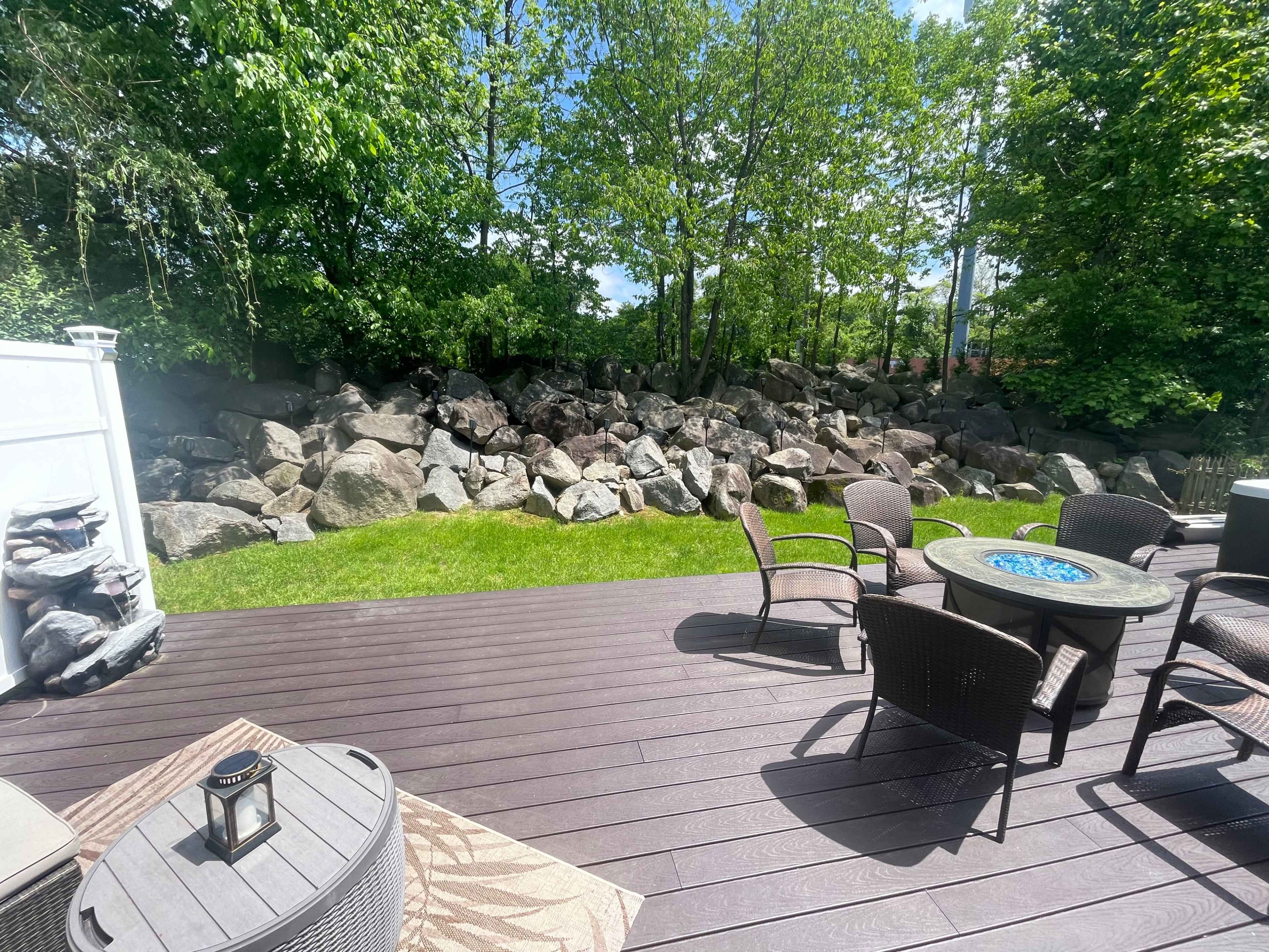 A wooden deck with outdoor furniture overlooks a grassy yard bordered by a large stone wall and trees.