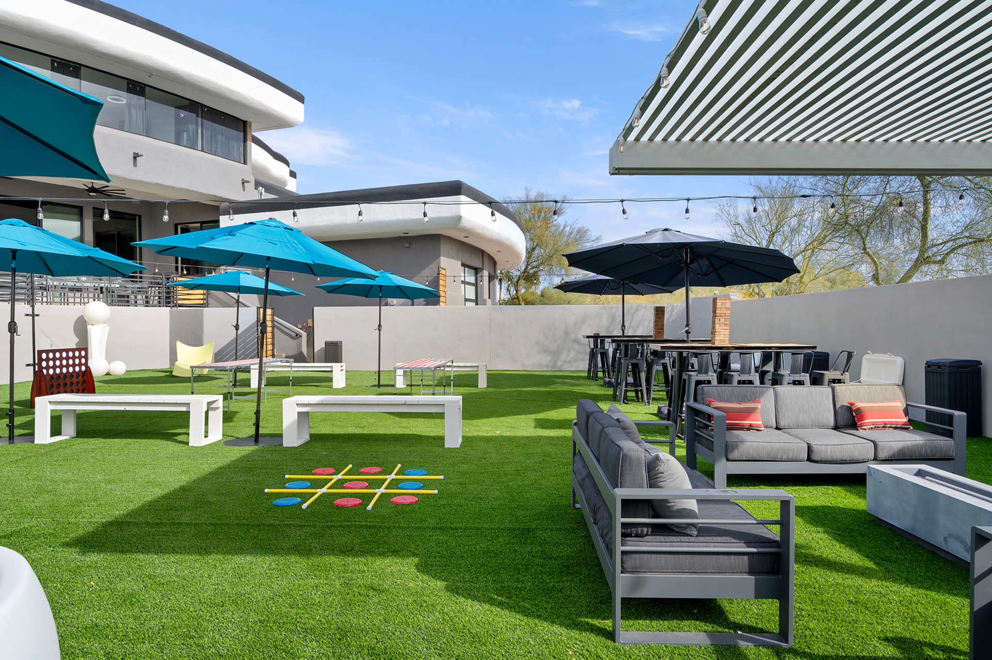 The image shows a modern outdoor space featuring artificial grass, seating areas with gray sofas, tables, blue umbrellas, and playful games.