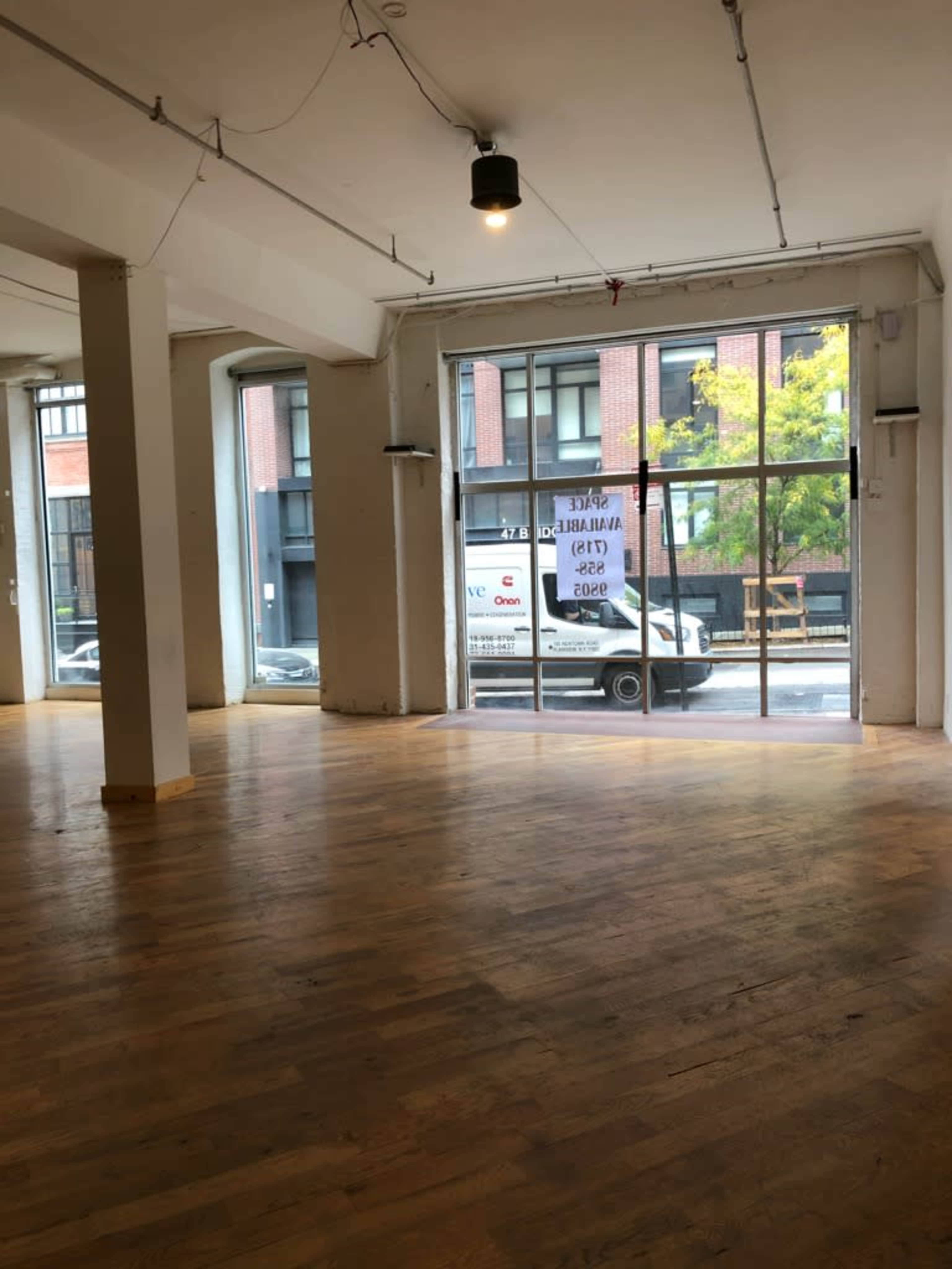 The image shows an empty interior space with wooden floors and large glass windows facing a street where a truck is parked outside.