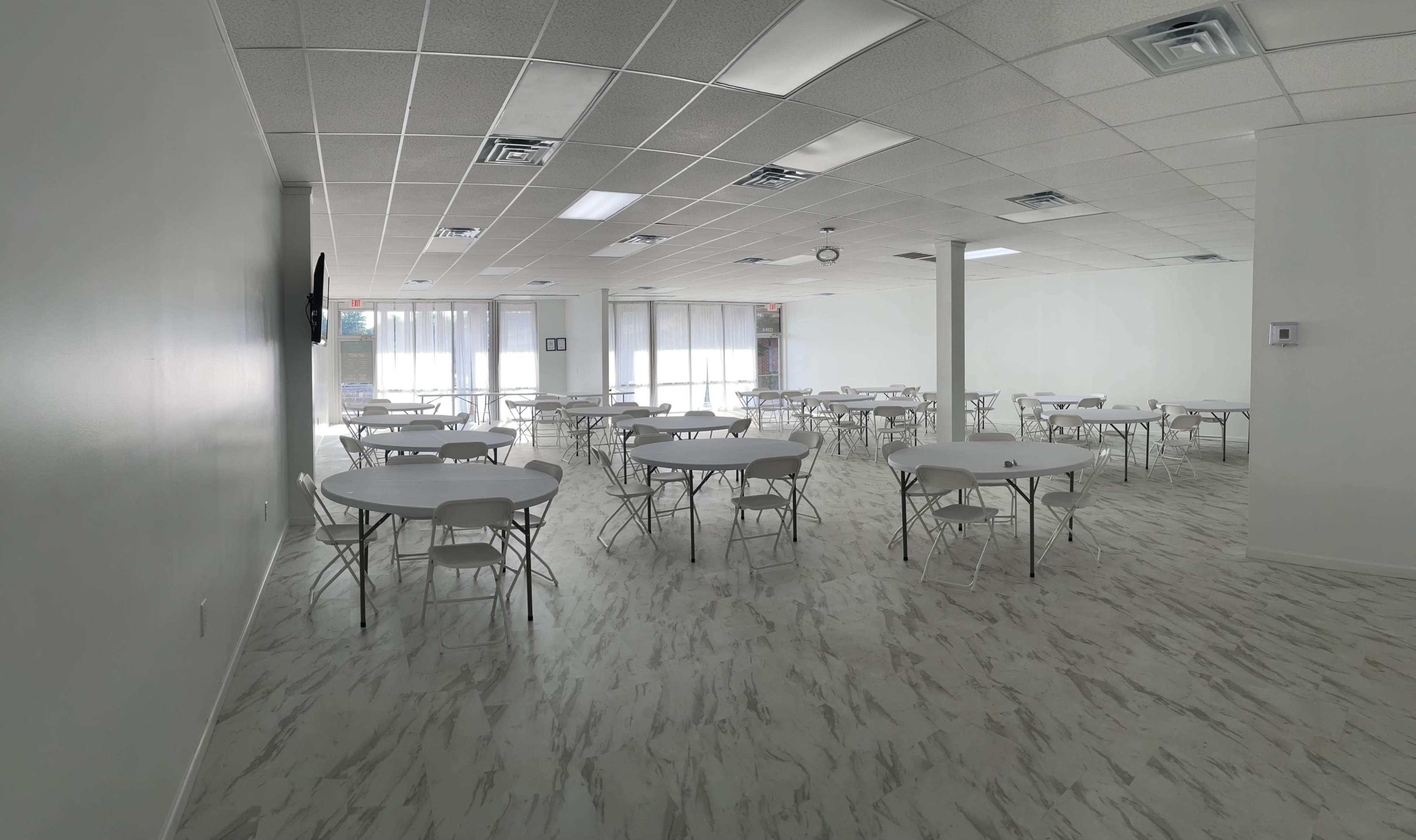 A spacious room with white walls and a patterned floor, featuring multiple round tables surrounded by chairs.