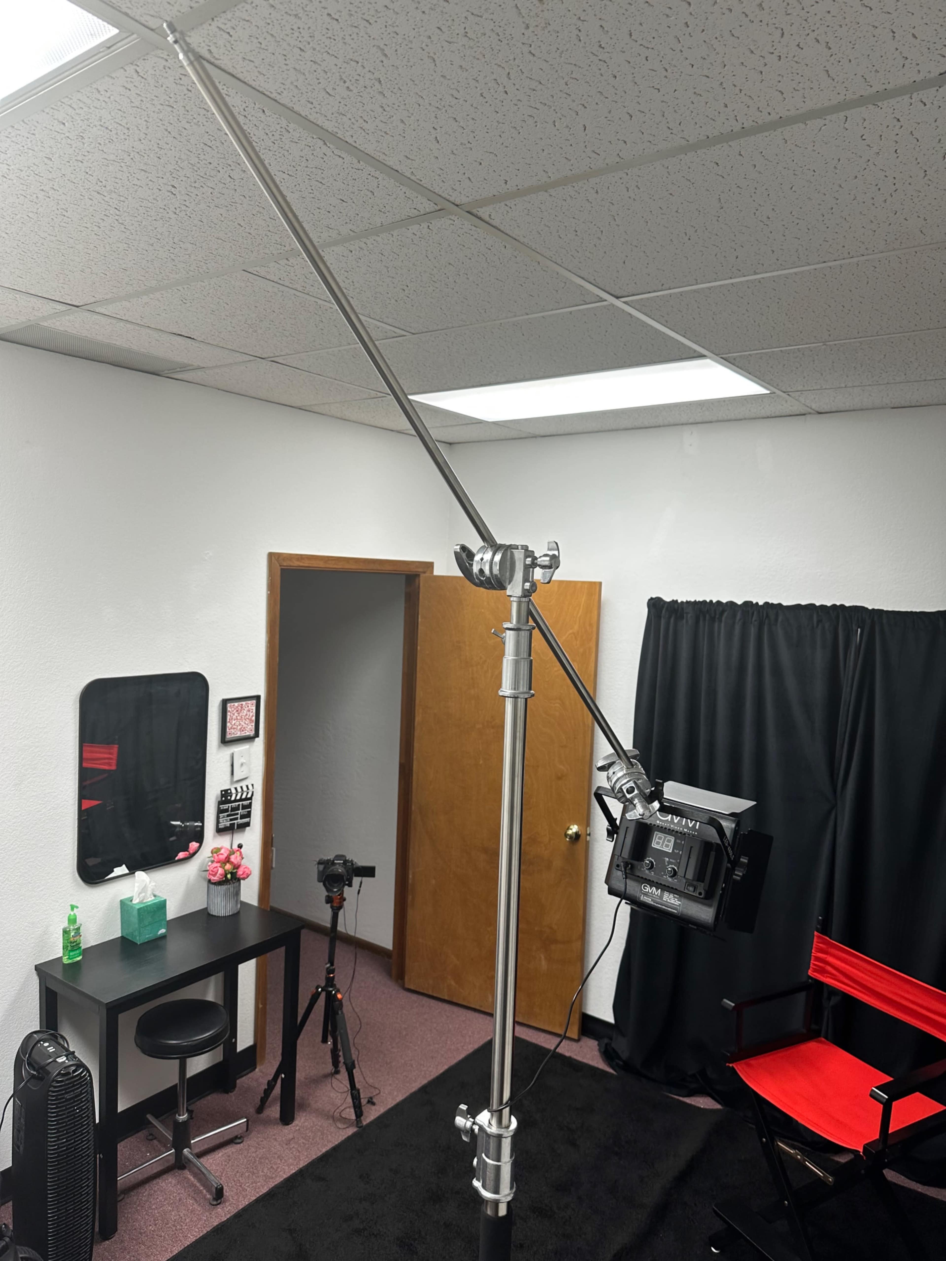 A lighting stand with an adjustable arm and a light fixture is positioned in a room featuring a desk, chair, and a camera setup.