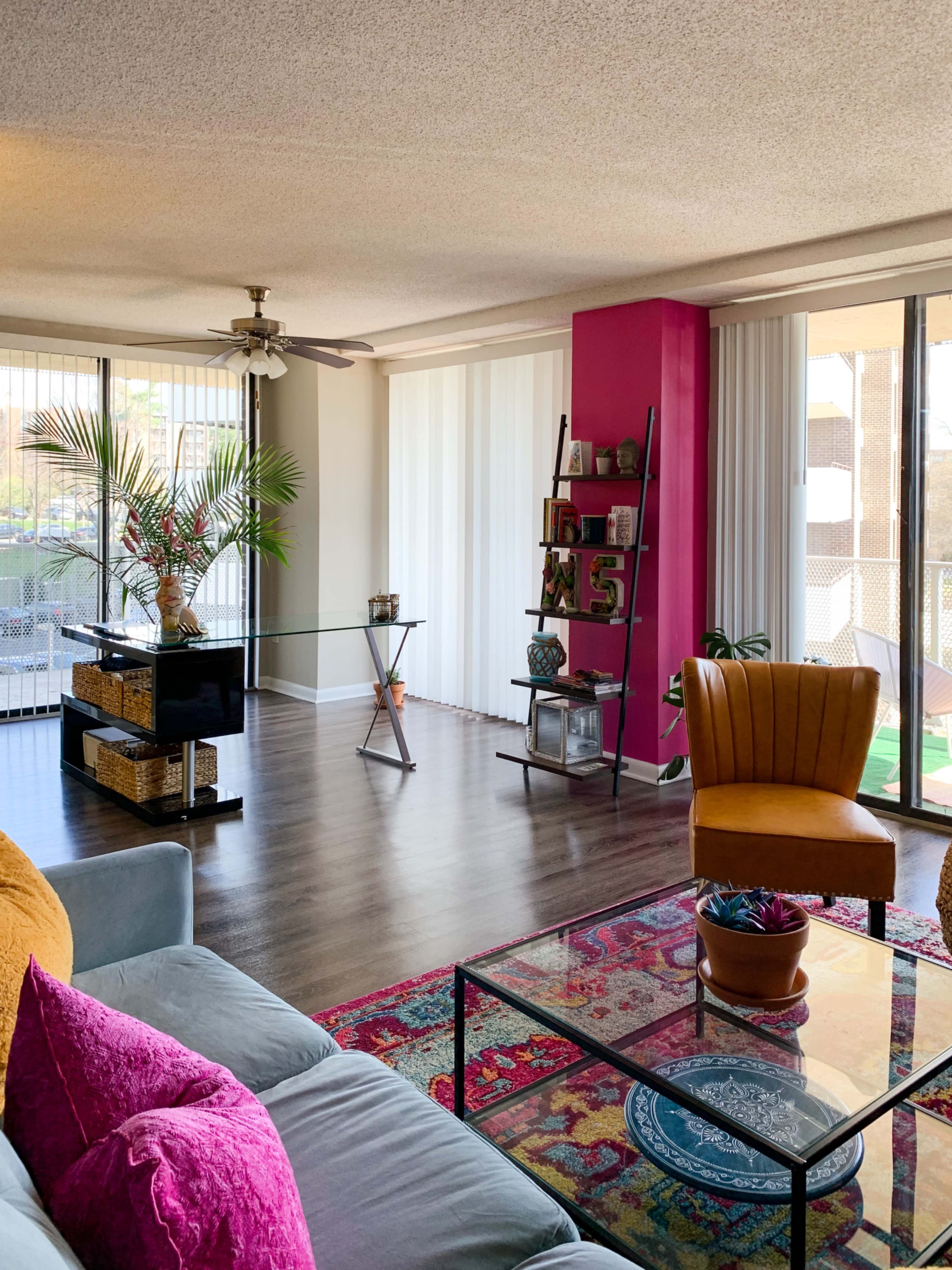 A bright living room featuring a mix of furniture, including a yellow armchair, a glass coffee table, and a bookshelf against a pink wall.