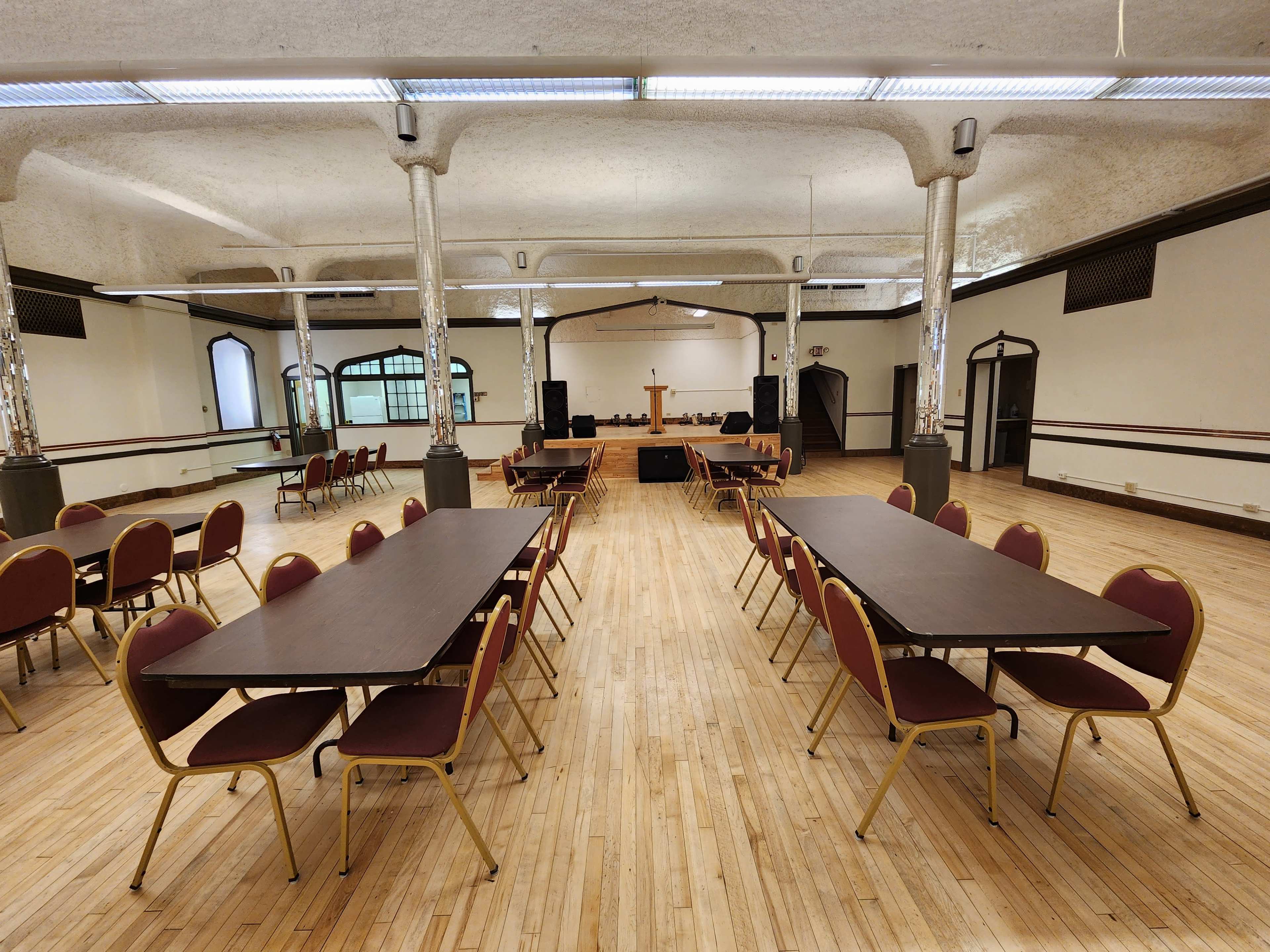 A spacious room with polished wooden floors features several tables arranged in rows and chairs positioned around them, facing a small stage at the front.