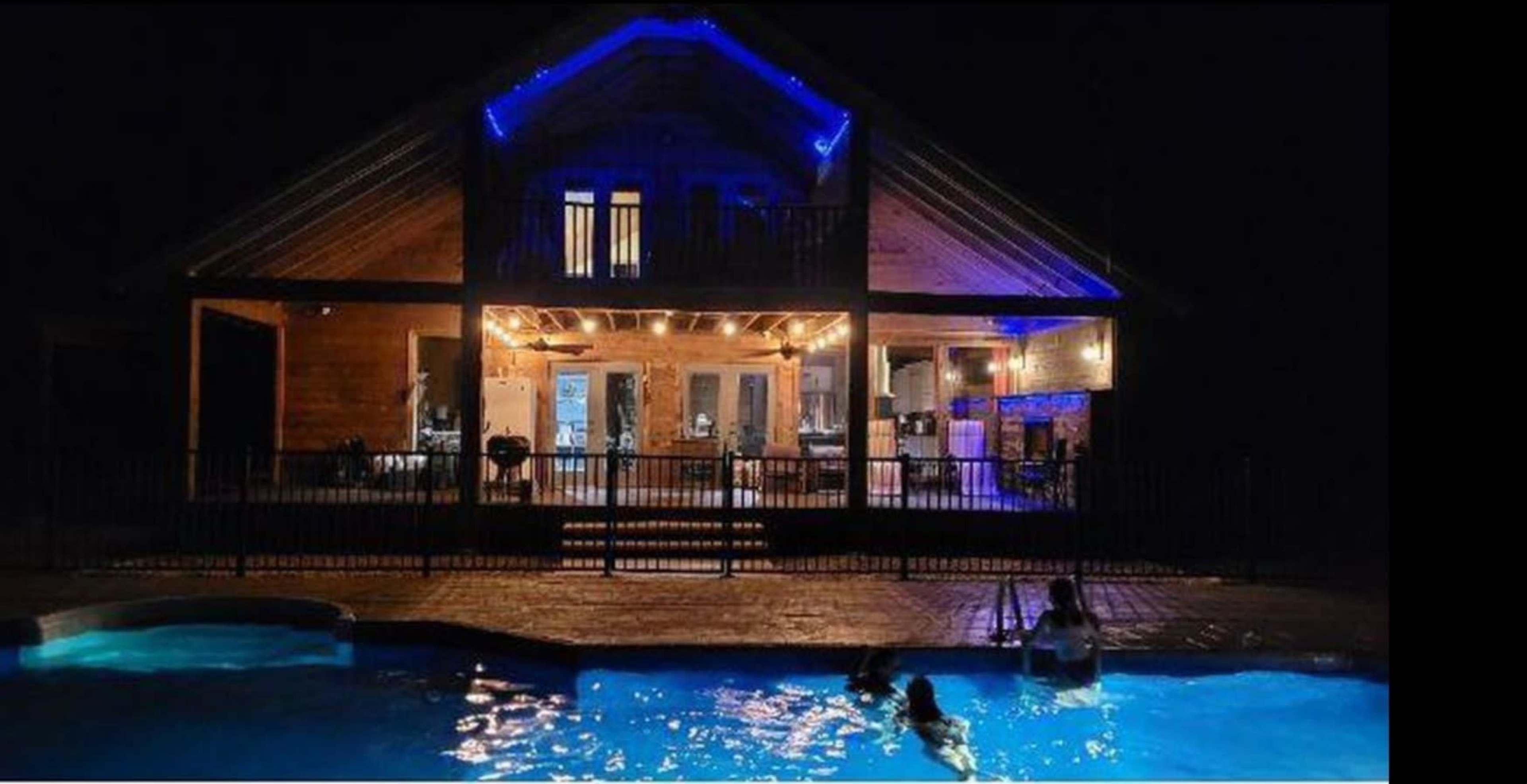 A large wooden house with illuminated features stands behind a swimming pool at night, with people swimming in the water.