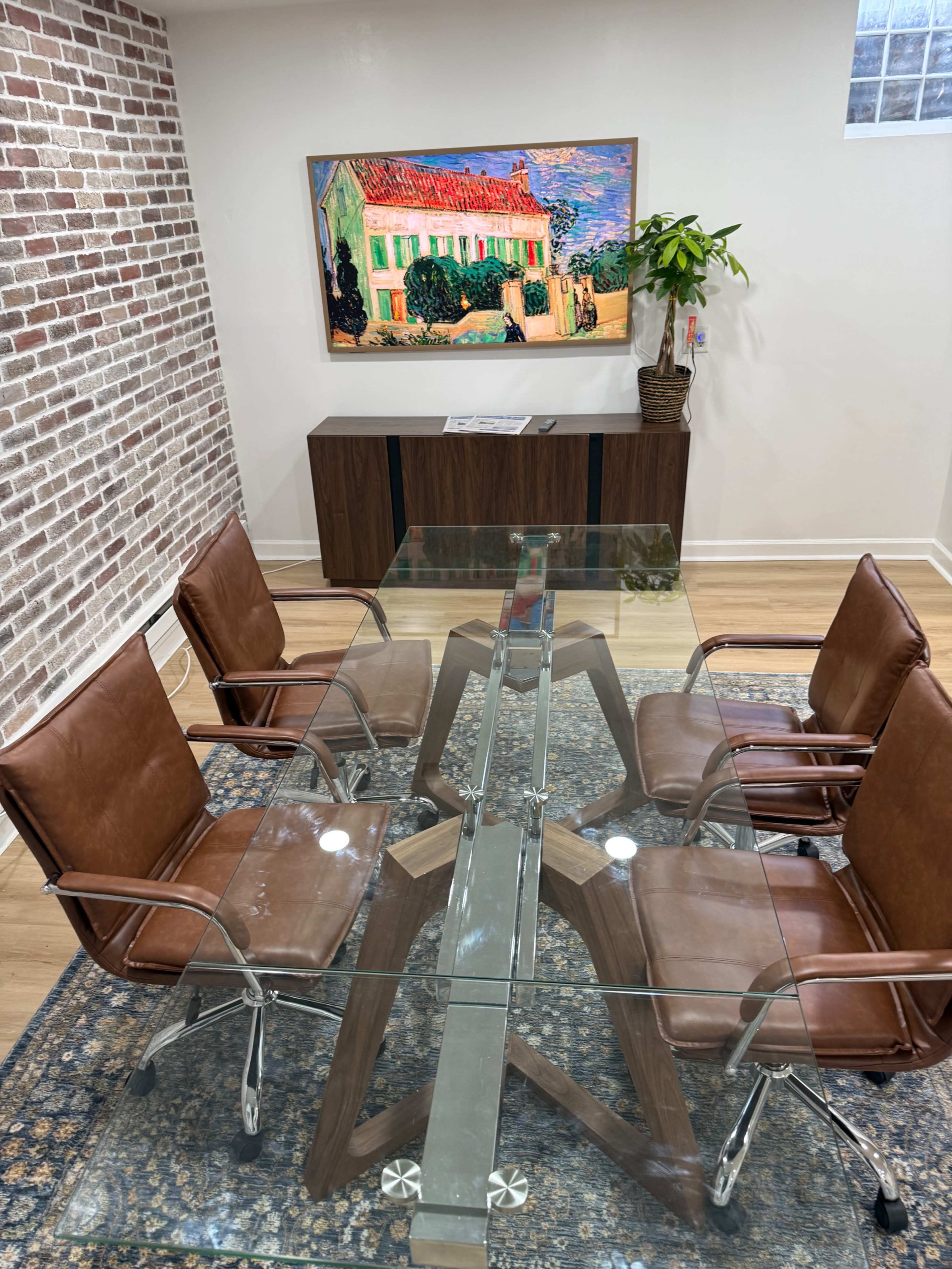 The image shows a modern conference room with a glass table surrounded by four brown leather chairs, featuring a brick wall and a colorful painting on the wall above a wooden cabinet.