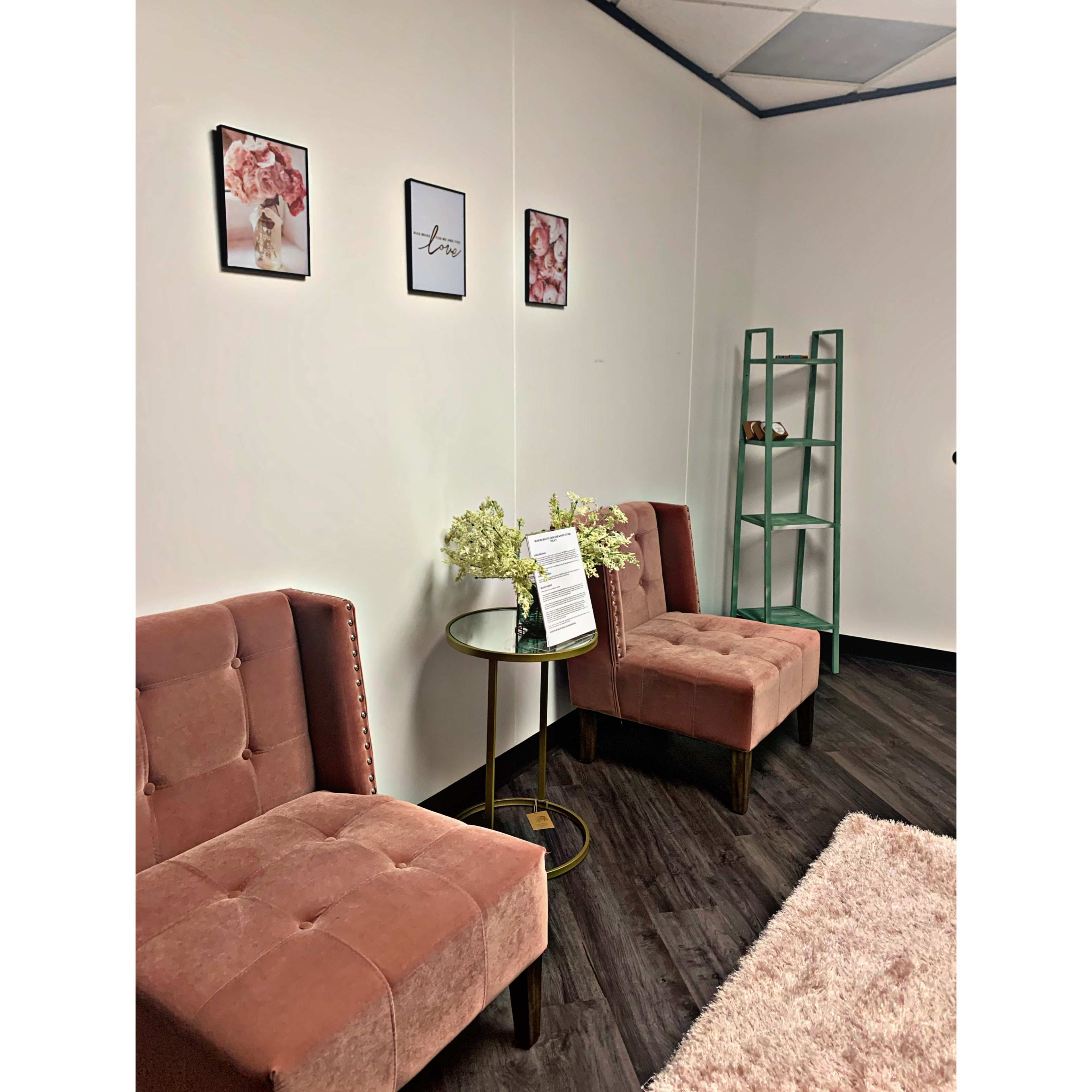 The image shows a small waiting area with two pink upholstered chairs, a round glass top table, a green shelving unit, and framed floral art on the walls.