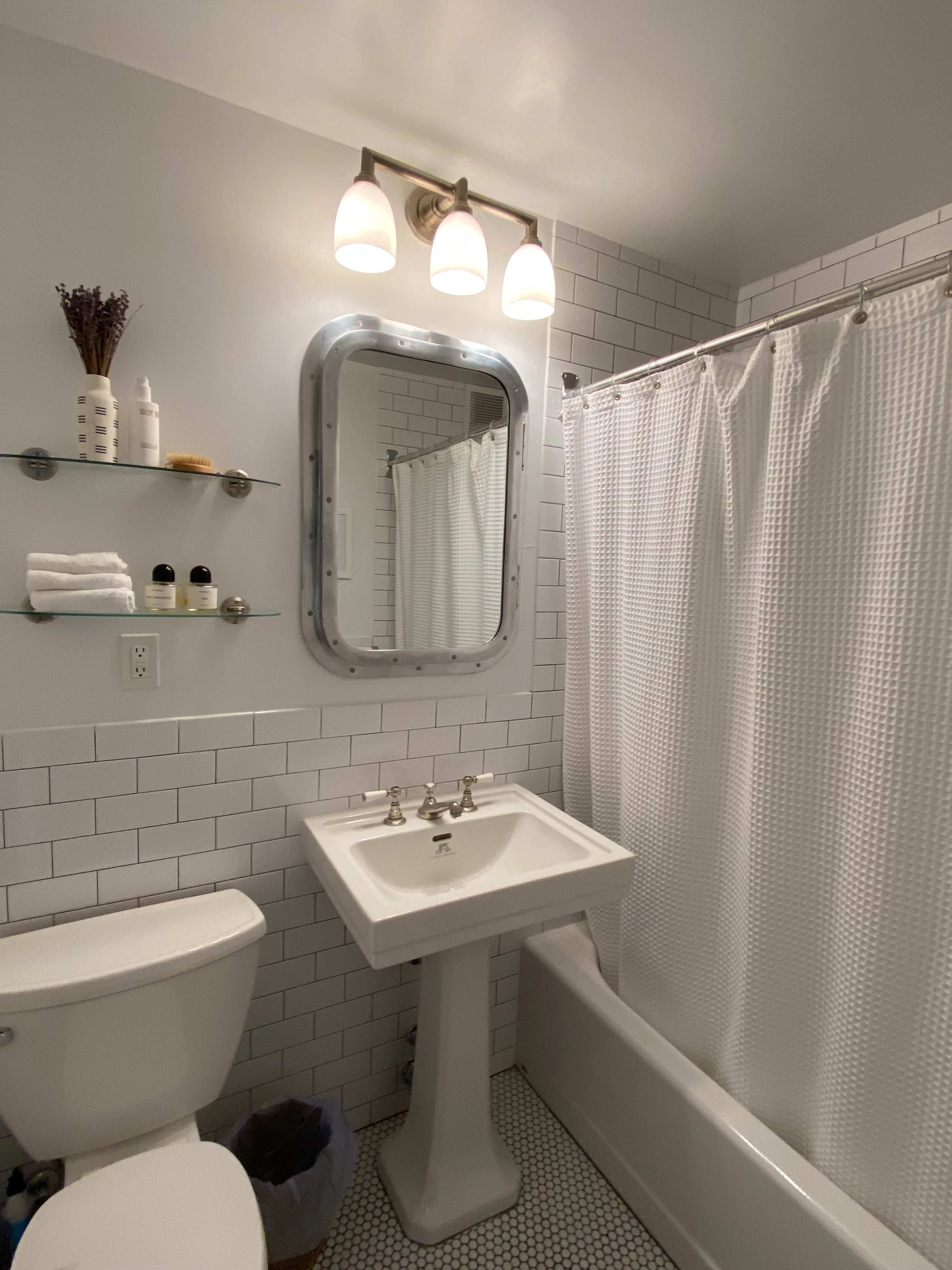 The image shows a small bathroom featuring a pedestal sink, a toilet, and a bathtub surrounded by white tile and a shower curtain.