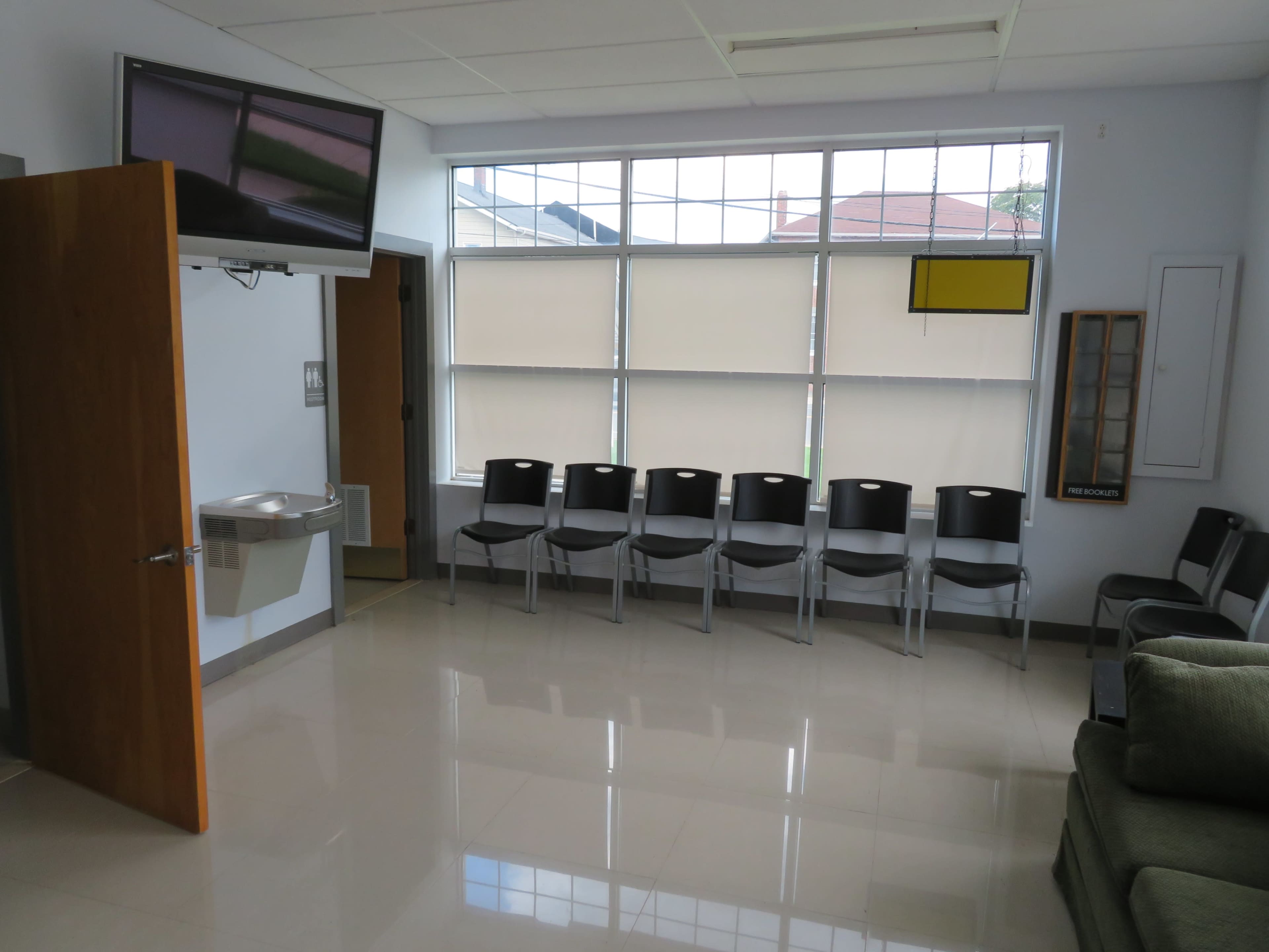 The image shows a waiting room with several black chairs lined up along a wall, a water fountain, a television mounted on the wall, and natural light coming through large windows.