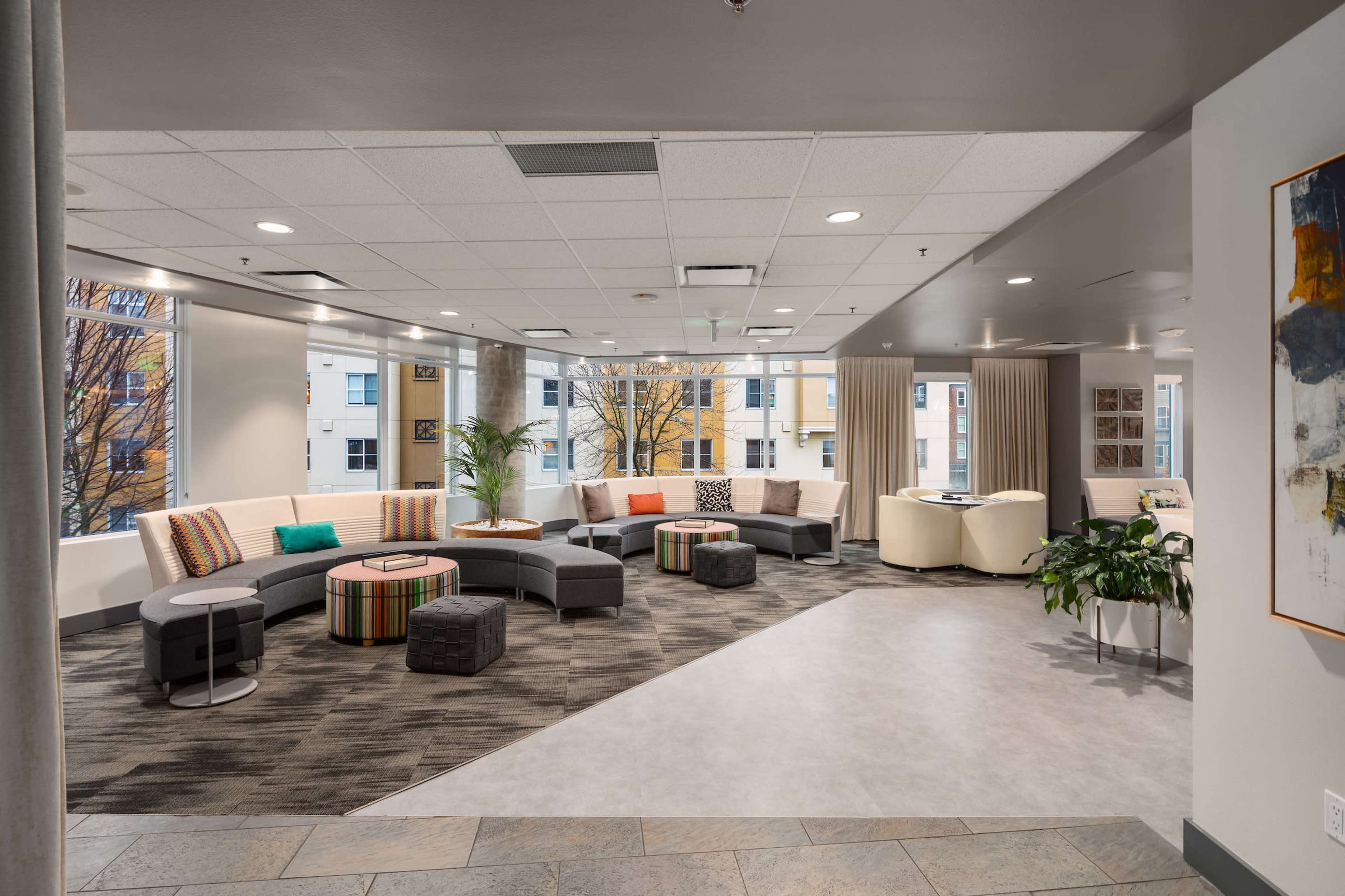 A modern lounge area with a curved sectional sofa, colorful cushions, small tables, and large windows allowing natural light to fill the space.