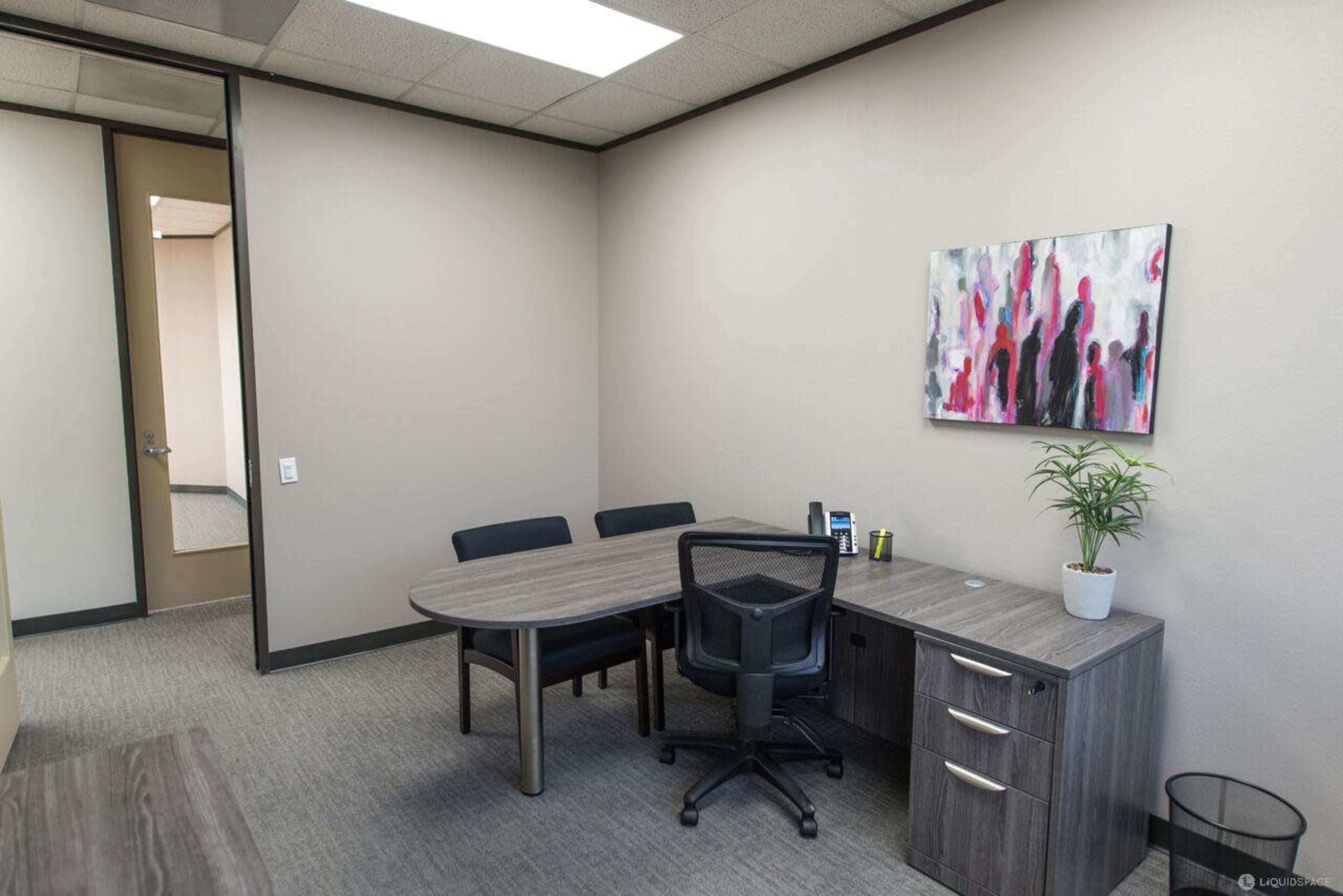 A modern office space with a round table and four chairs, a desk with drawers, and a plant in a pot against a plain wall.