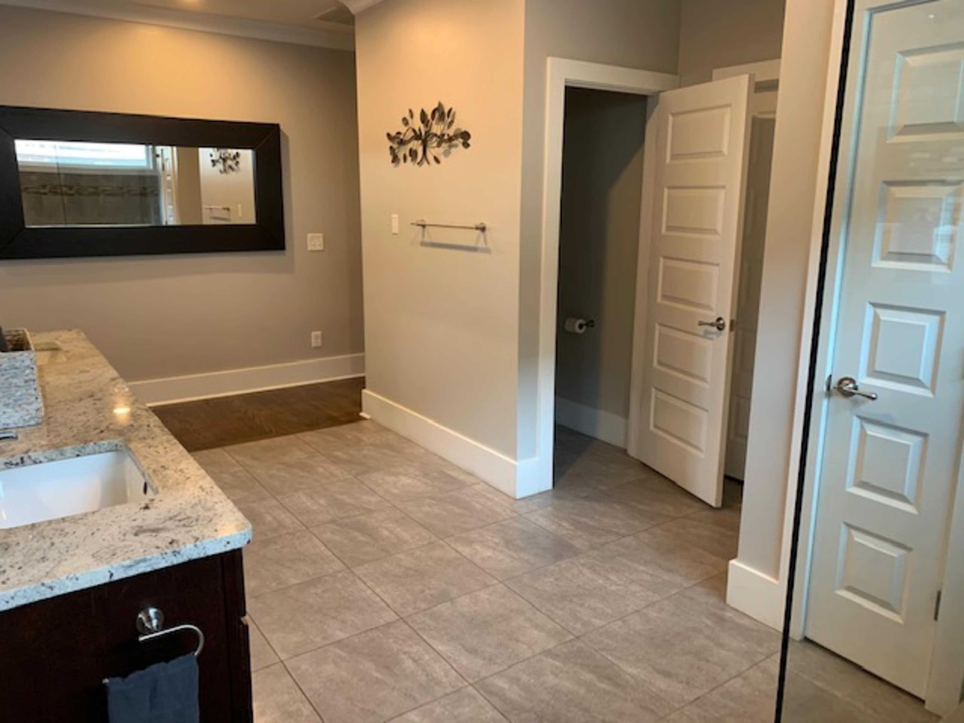 A spacious bathroom featuring a gray tiled floor, a granite countertop vanity with a mirror, and a doorway leading to a toilet area.