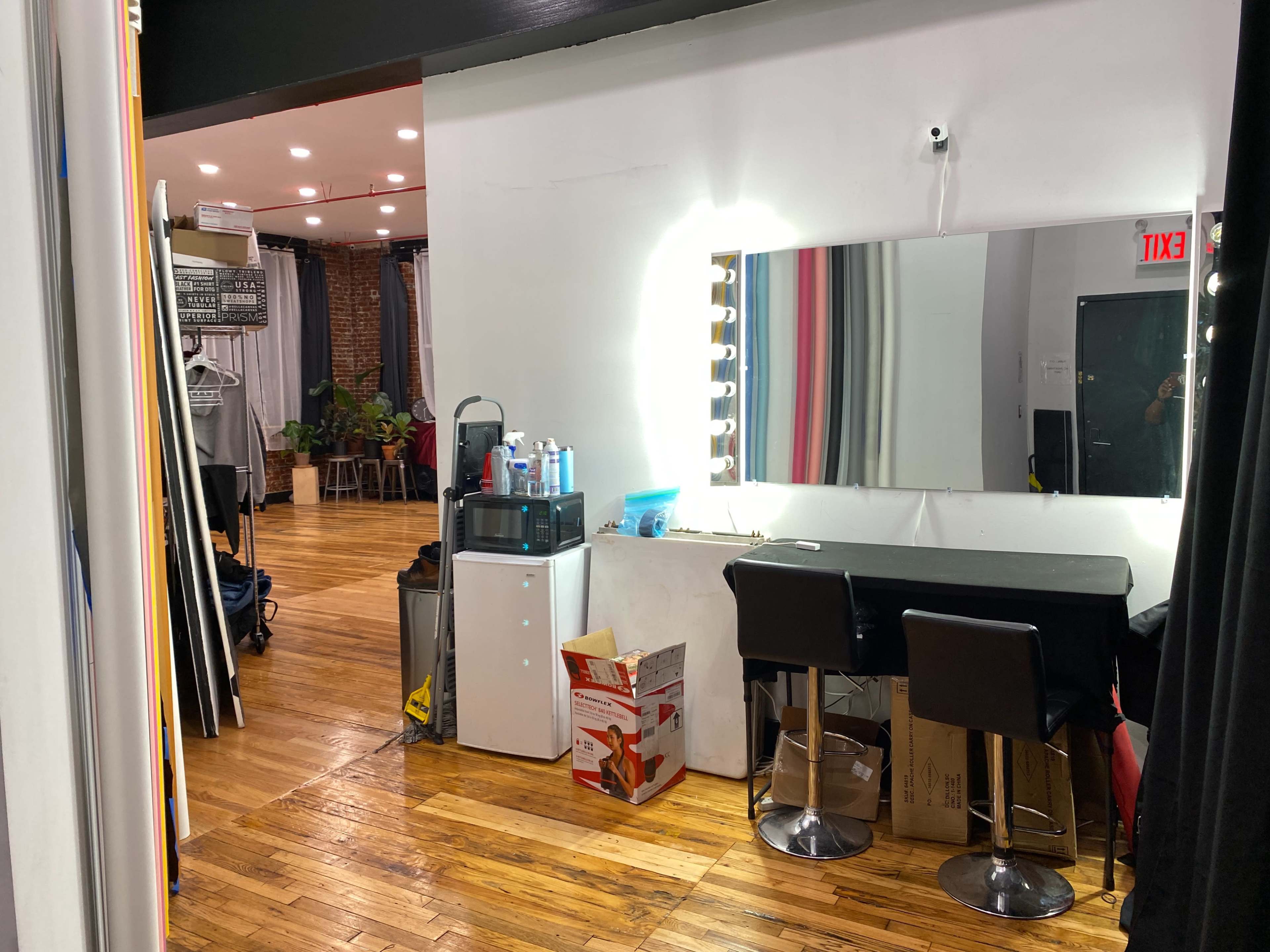 The image shows a room with a small kitchen area on the left, a large mirror with lights above it on the right, and a table with black chairs in front of the mirror.