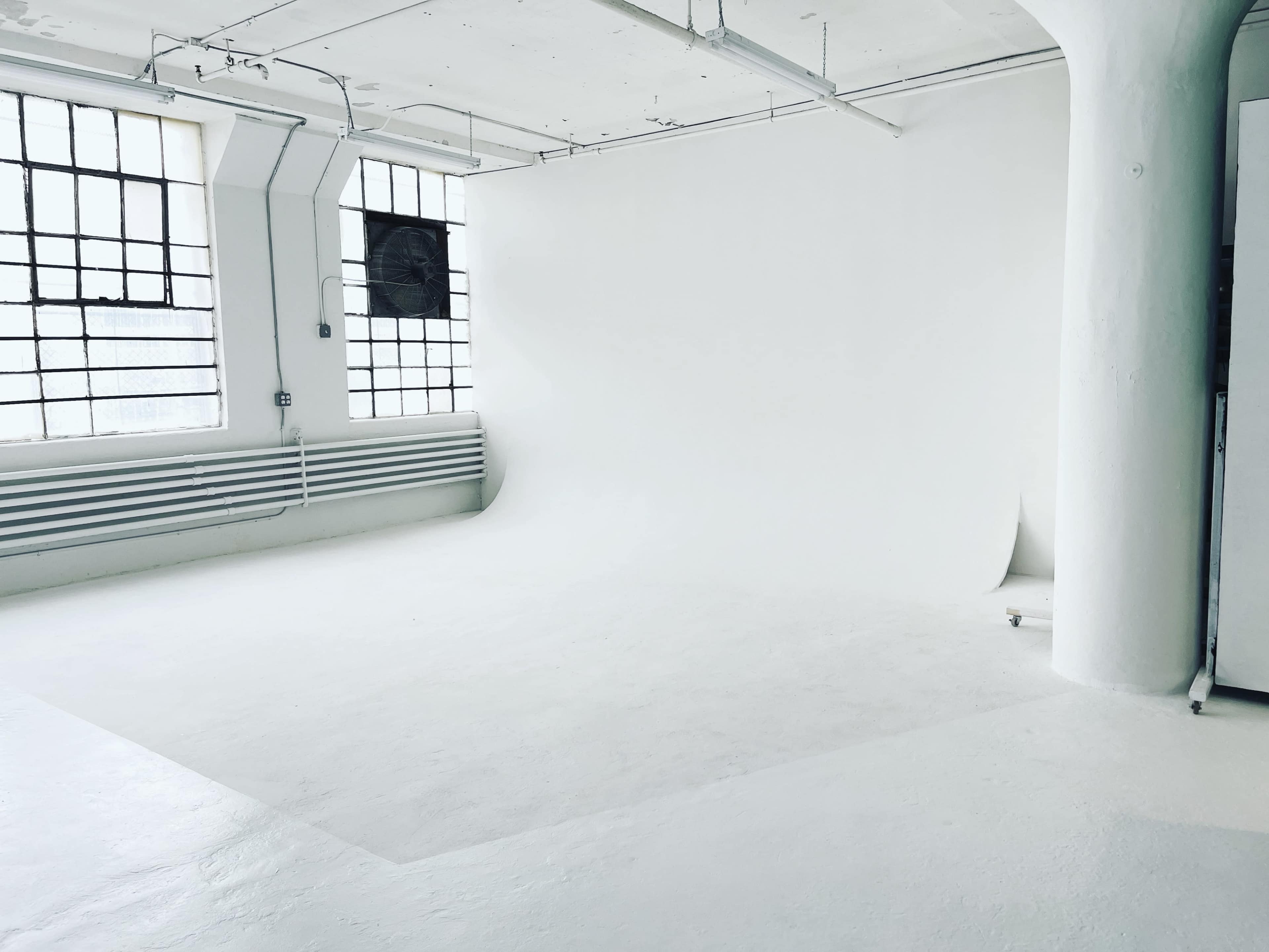 A spacious, empty studio featuring white walls, a white backdrop, and large windows.