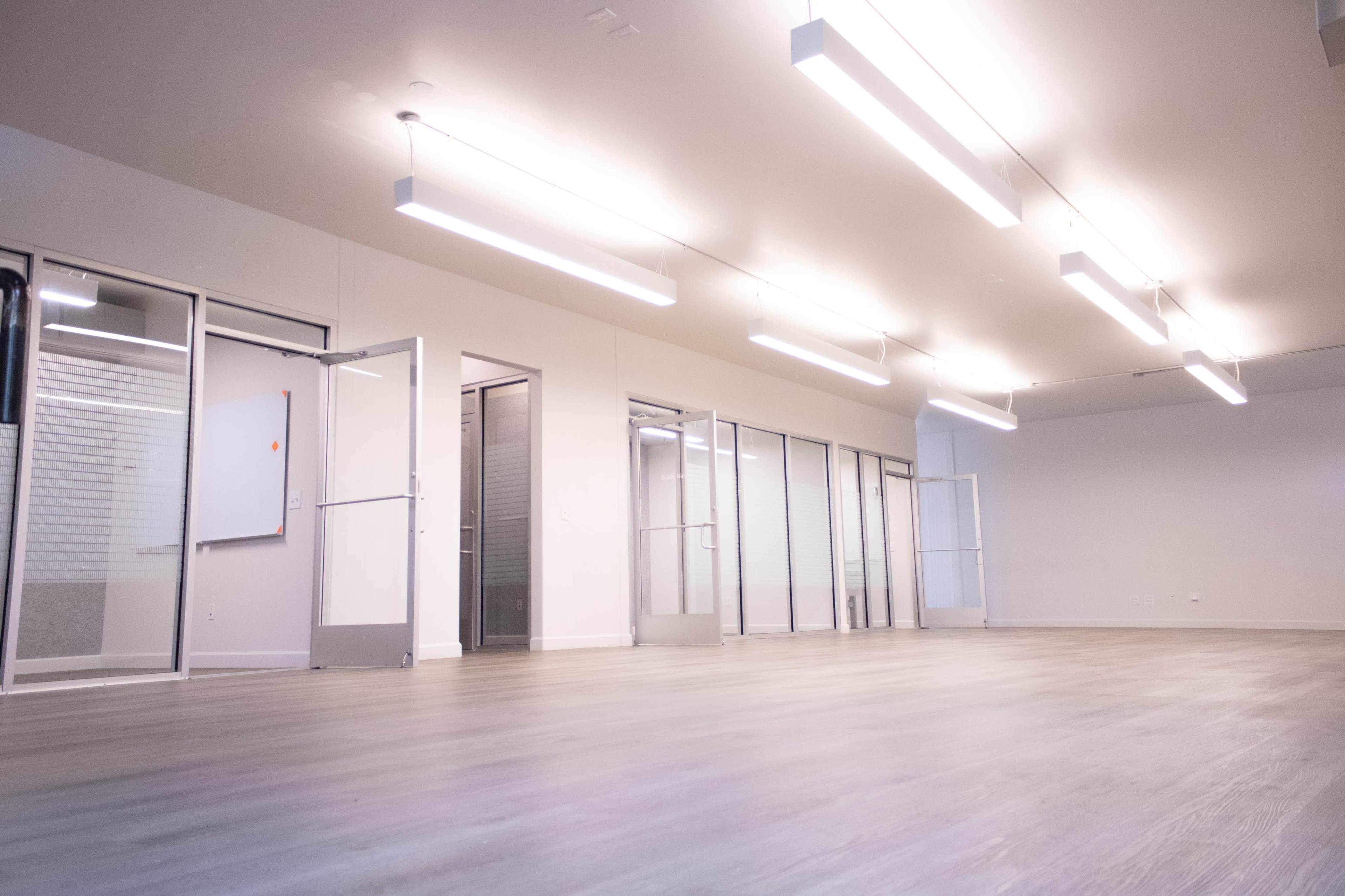 The image shows a spacious, empty office with large windows and modern lighting fixtures on the ceiling.