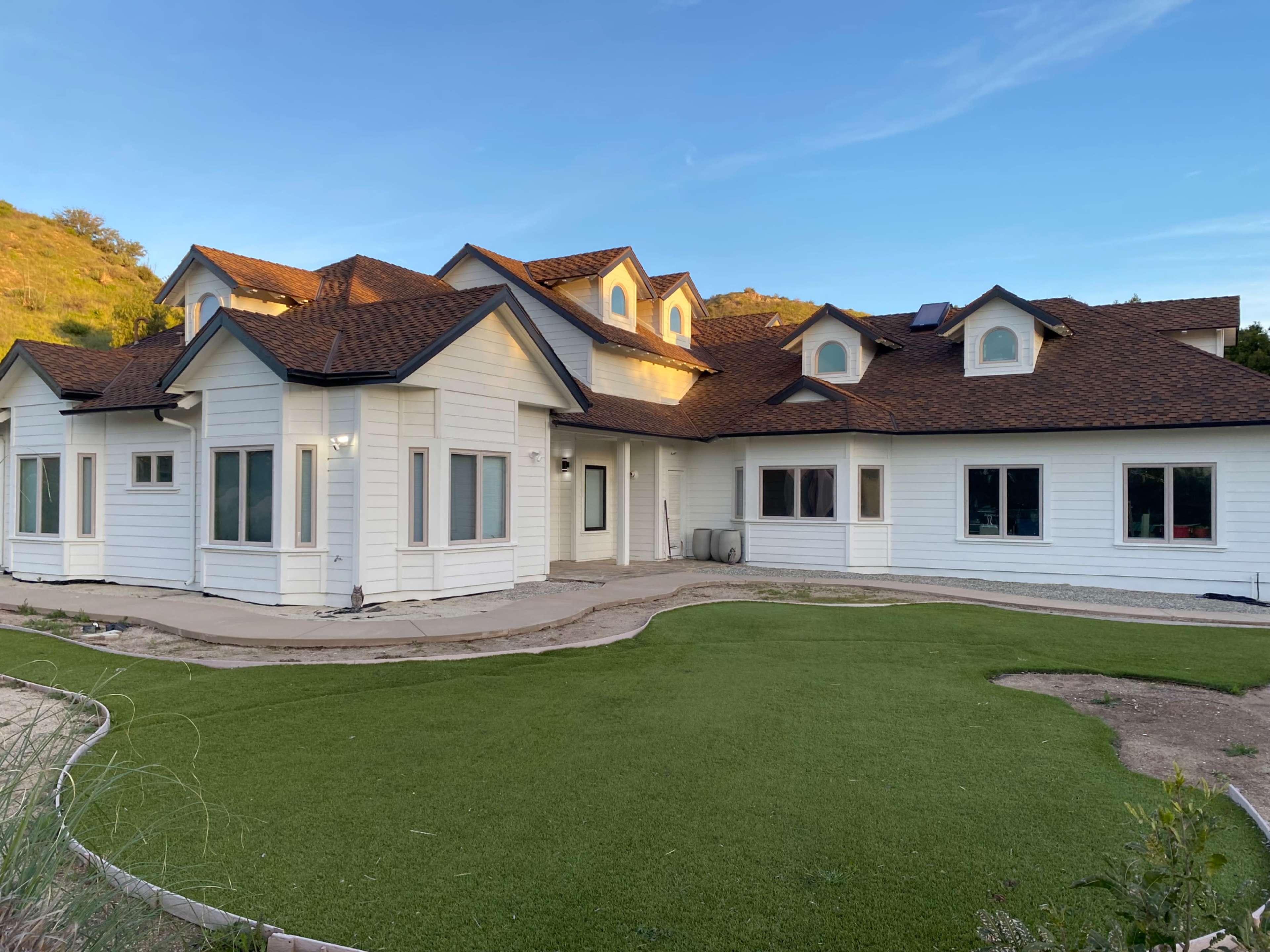 The image shows a large white house with a brown roof and multiple gabled sections, surrounded by a green lawn.