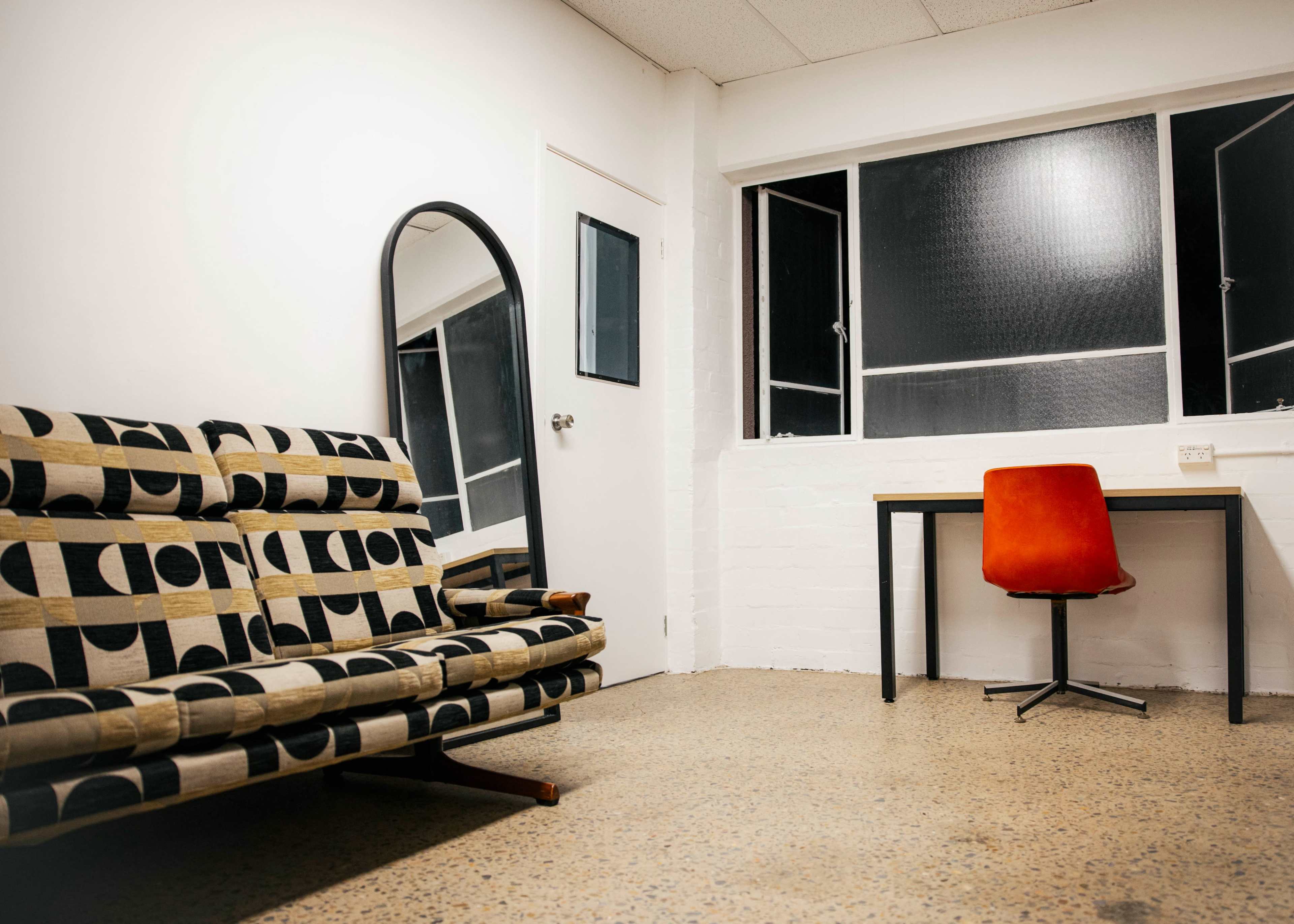 A minimalist room features a patterned sofa, a small wooden table, and an orange chair, with a large mirror against one wall and a window on another.