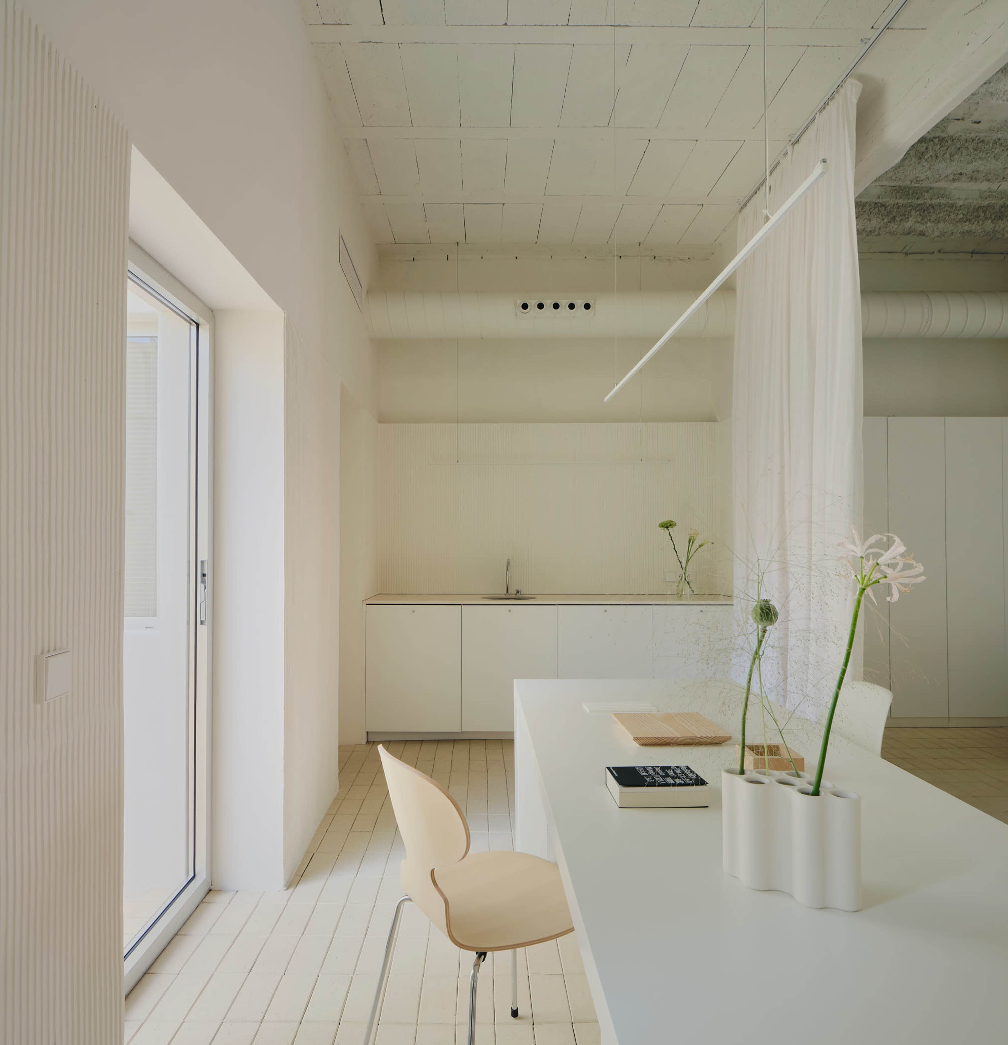 A minimalist kitchen space features a simple white table with a chair, natural light streaming through a large window, and a clean, uncluttered countertop.