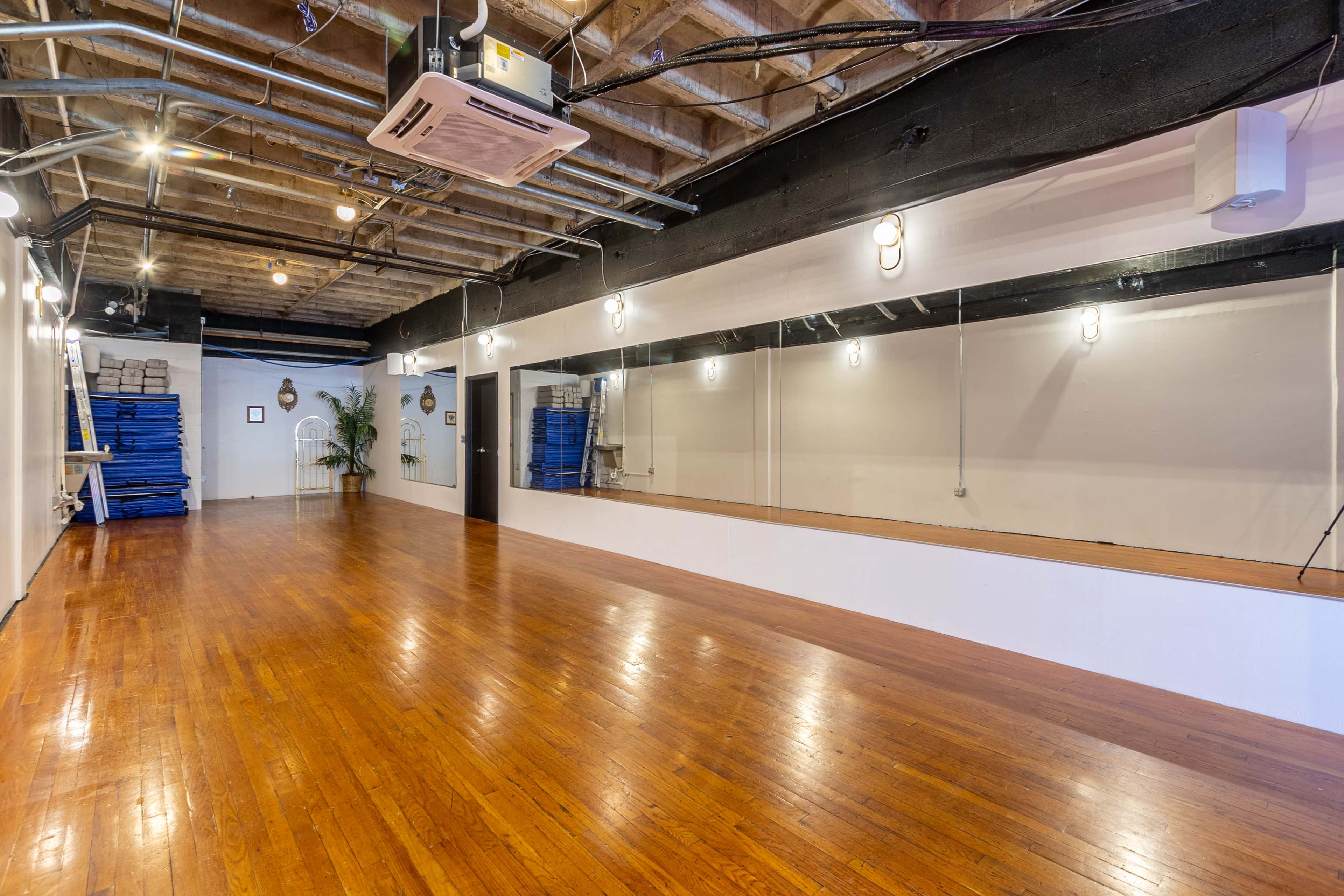 An empty studio space with wooden floors, a large mirror on one wall, and exposed beams on the ceiling.