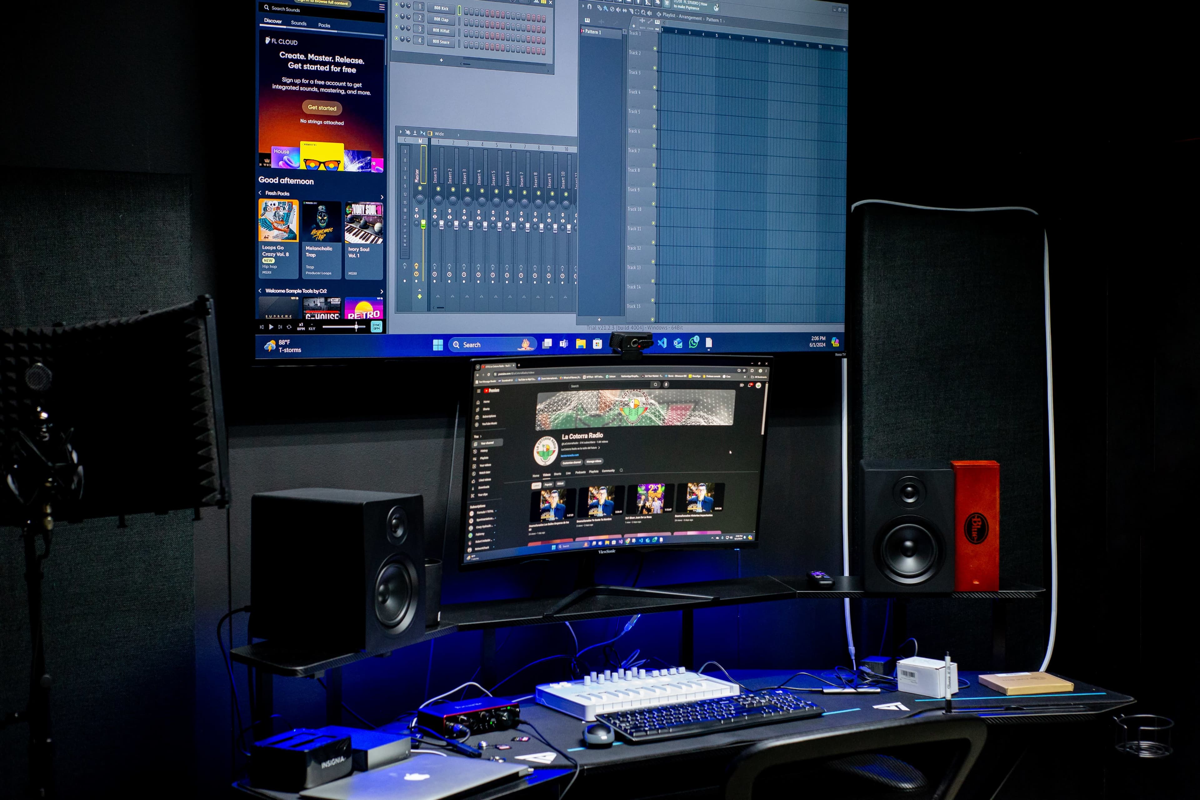 A modern music production studio with a large monitor displaying audio software, a smaller monitor with a video platform, and audio speakers on a desk equipped with various musical instruments and a MIDI keyboard.