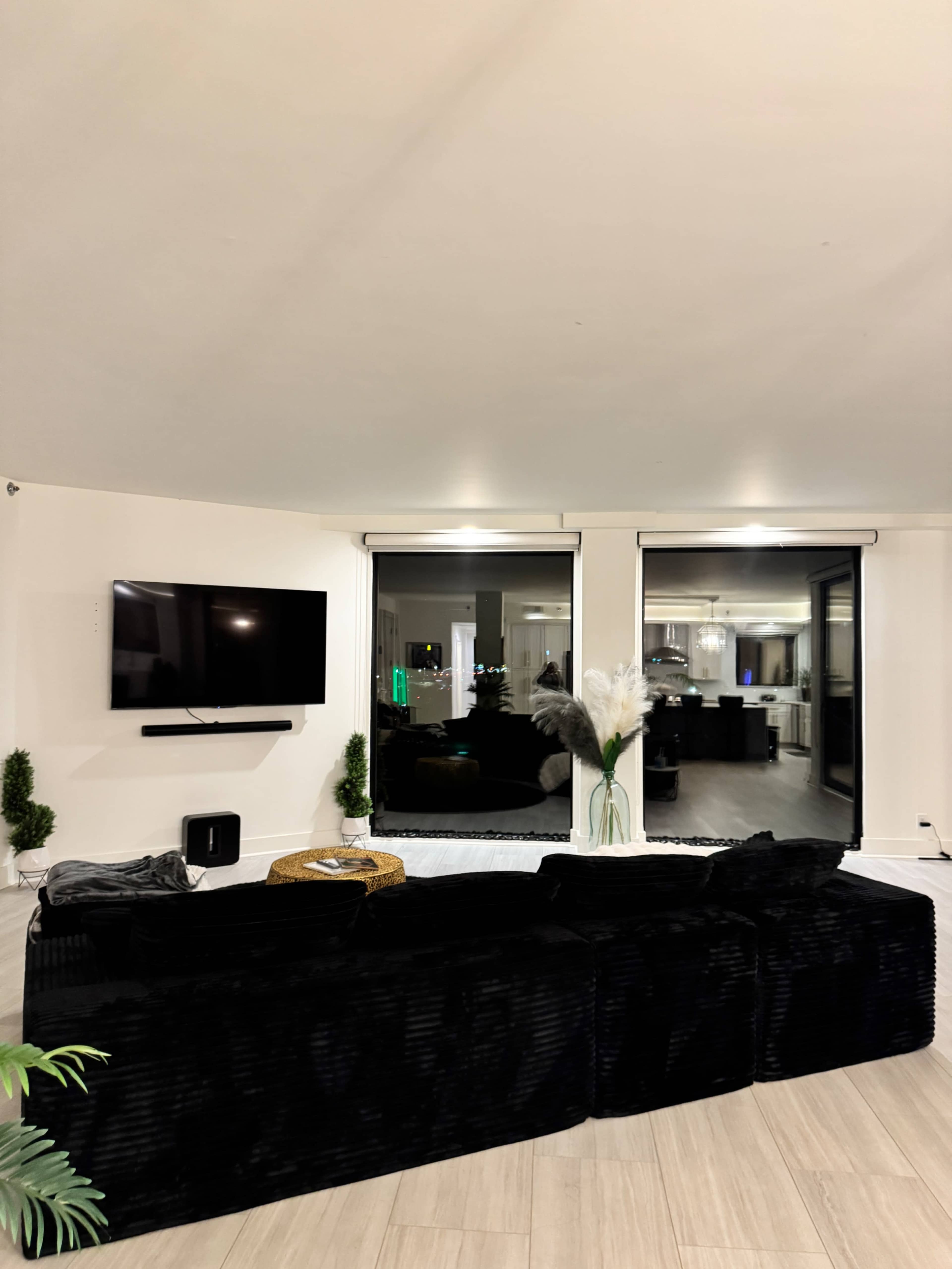 The image shows a modern living room featuring a black sectional sofa, a wall-mounted television, and large windows revealing a glimpse of another room.