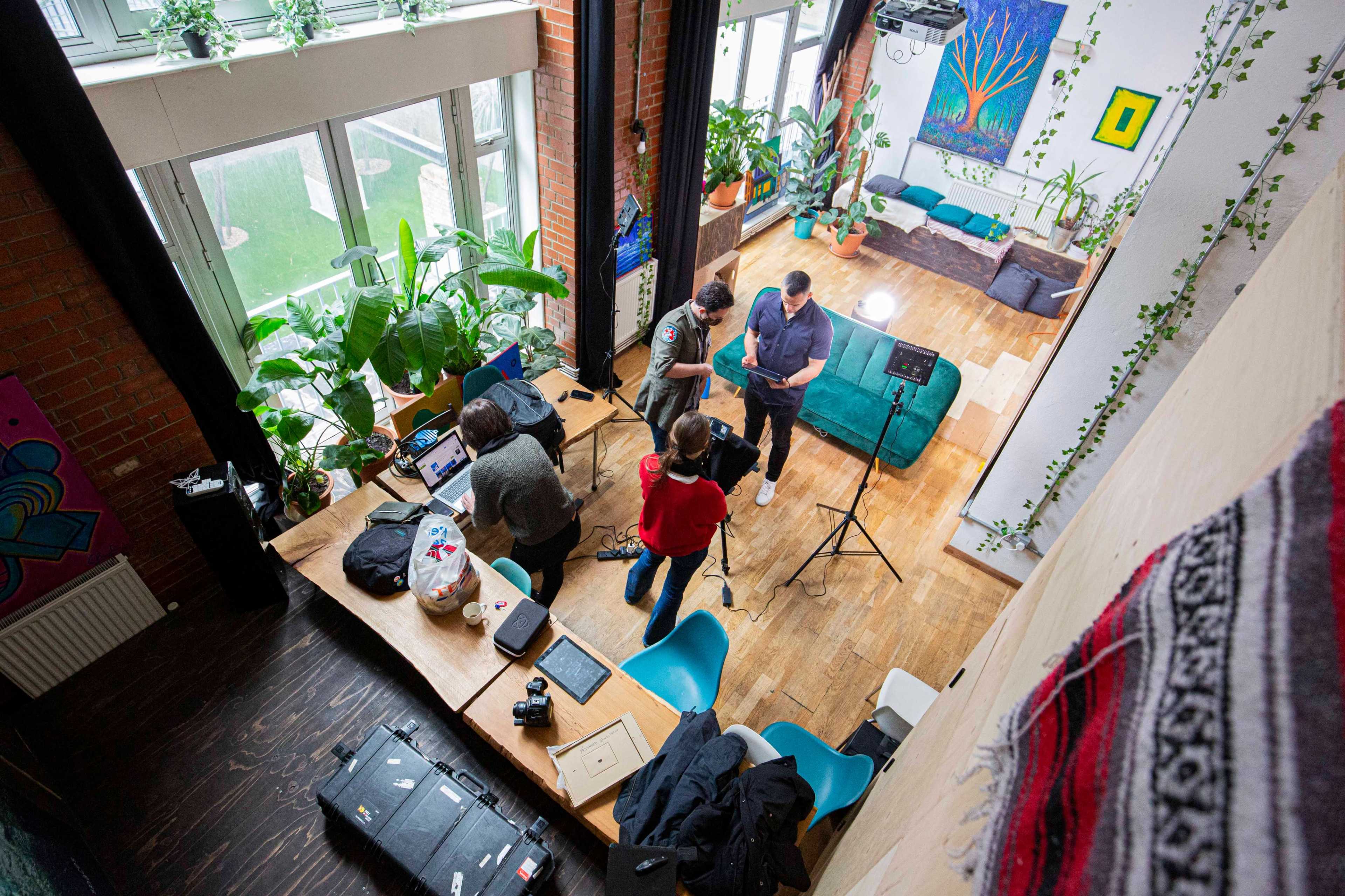 A group of four people set up equipment around a large wooden table in a brightly lit indoor space filled with plants and colorful artwork.