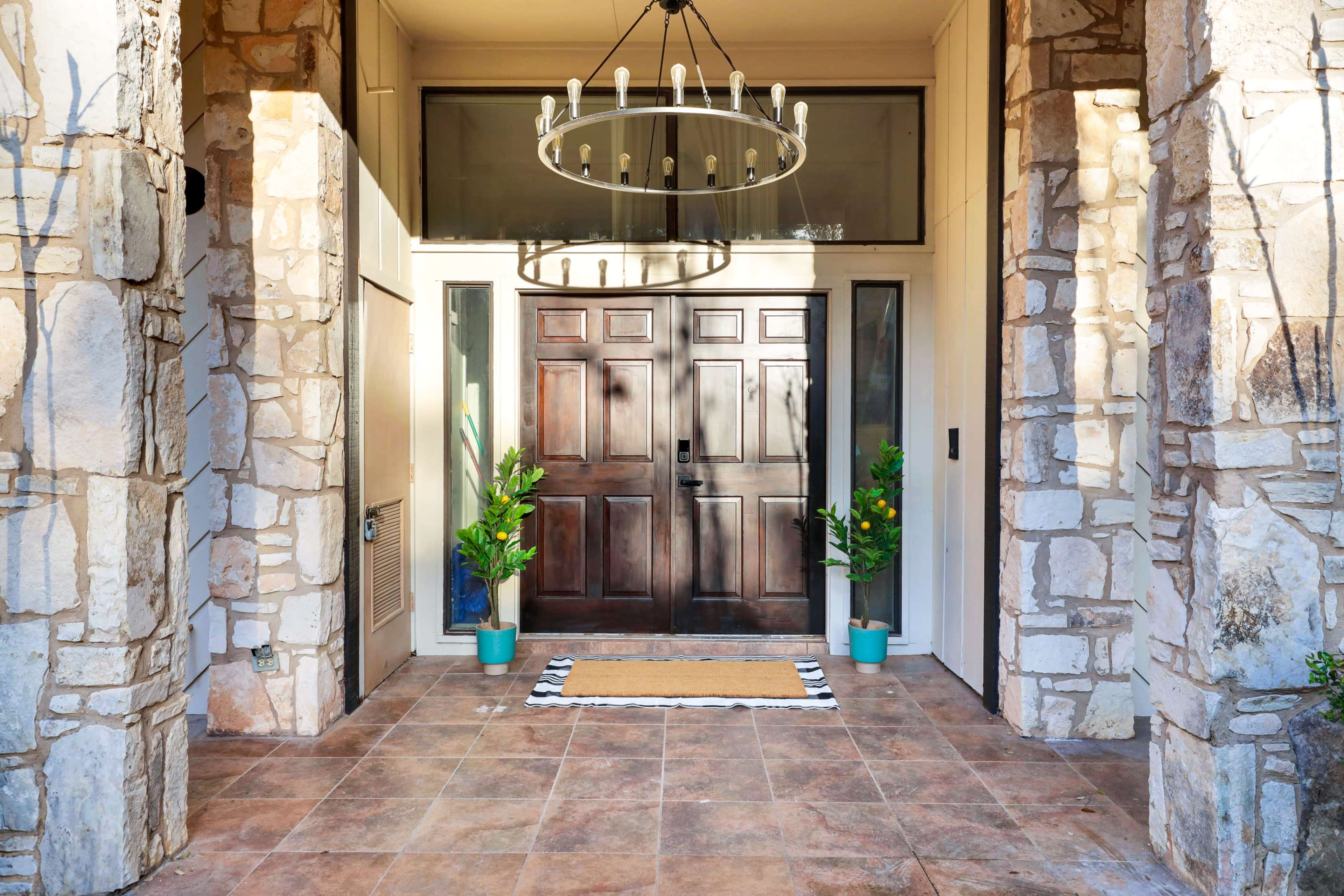 The entrance features double wooden doors flanked by potted plants and a chandelier overhead, with a tiled floor and a welcome mat.