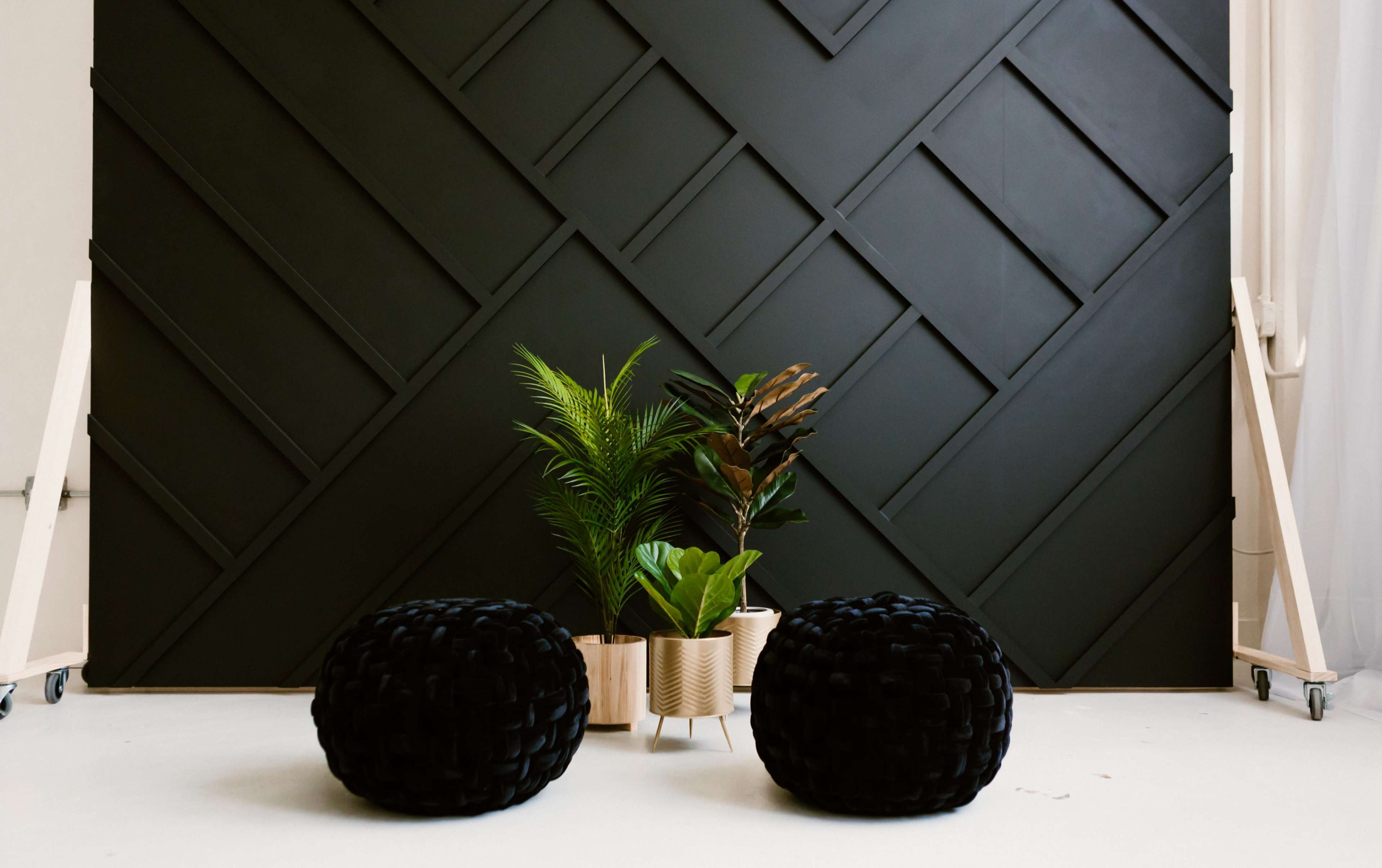 Two black poufs in front of a textured black wall, with a pair of plants in neutral-colored planters positioned between them.