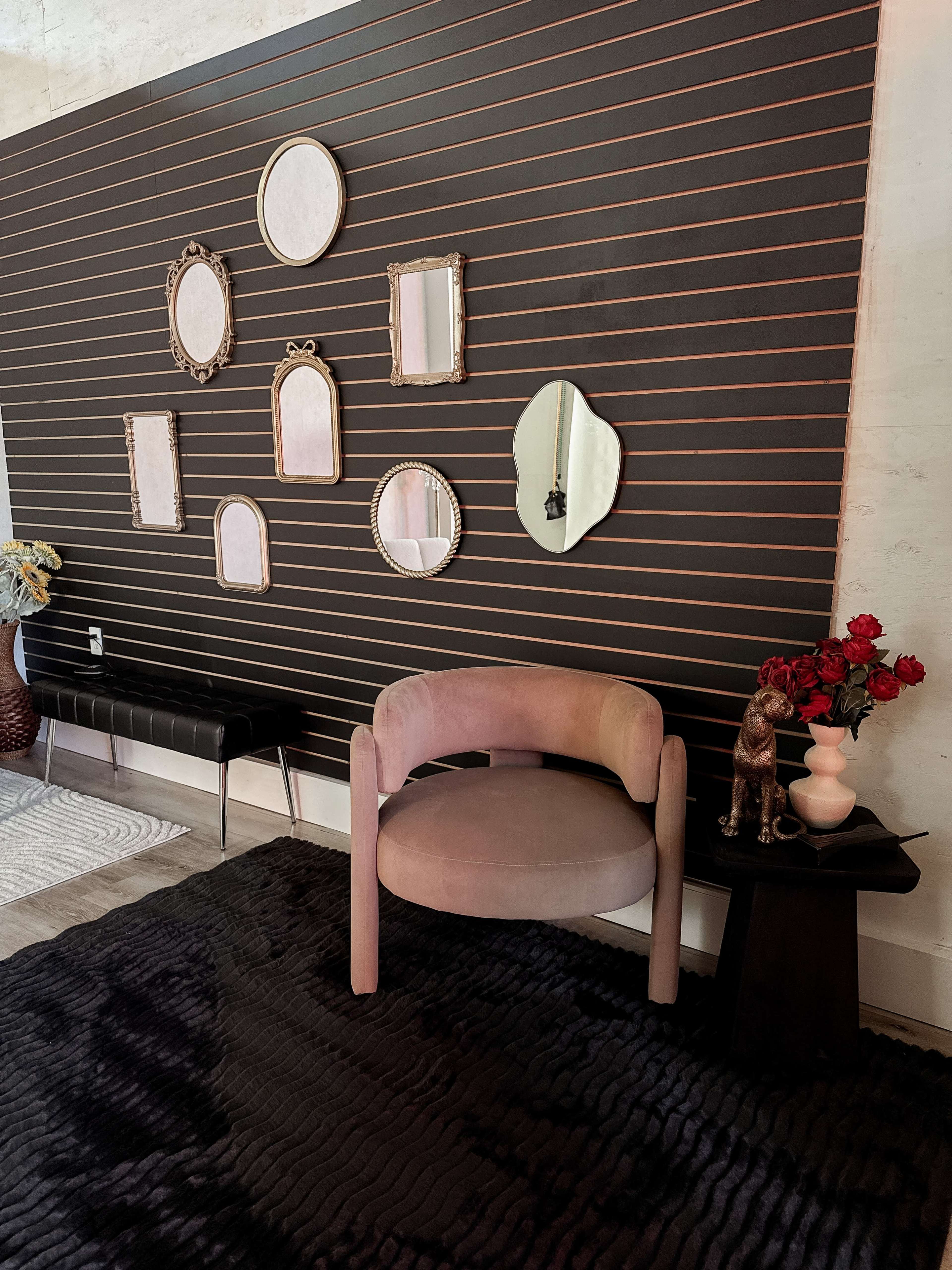 The image features a modern sitting area with a curved chair, a small table beside it, and several decorative mirrors arranged on a dark slat wall.
