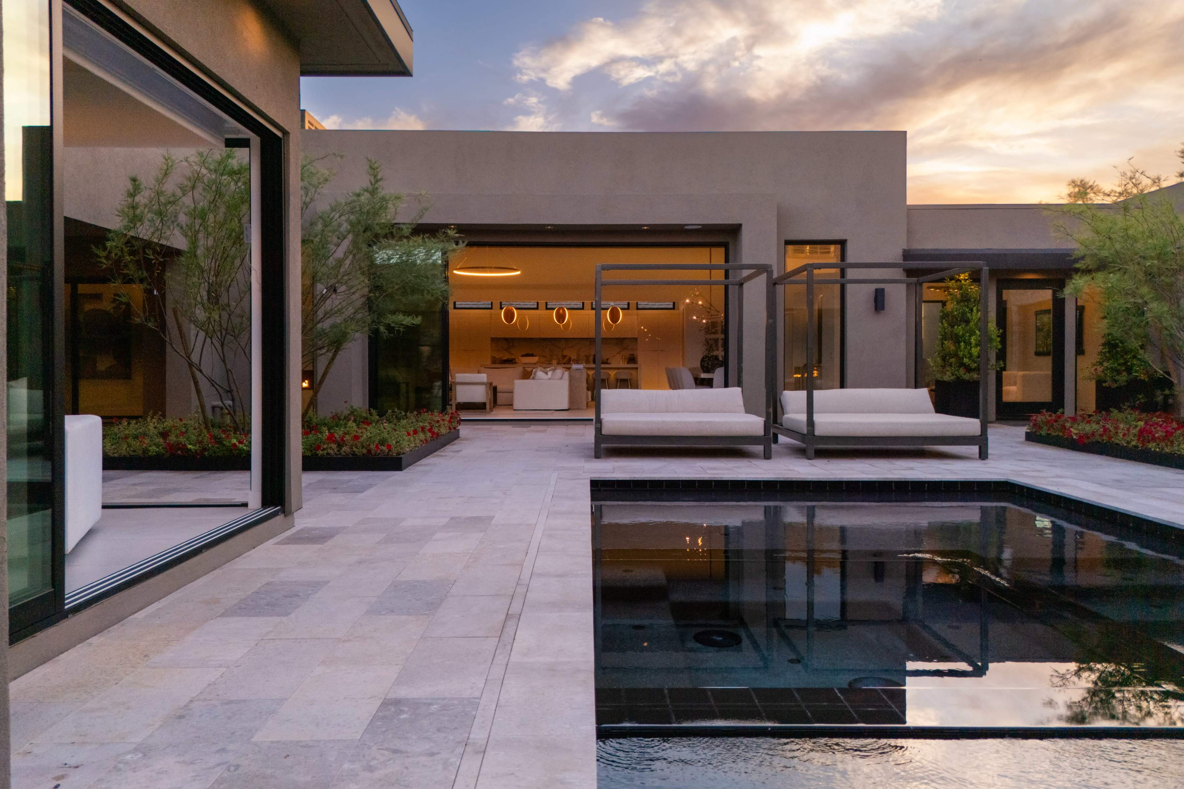 The image shows a modern outdoor area with a small pool, two lounge chairs, and an open interior space visible through large glass doors, set against a vibrant sunset sky.