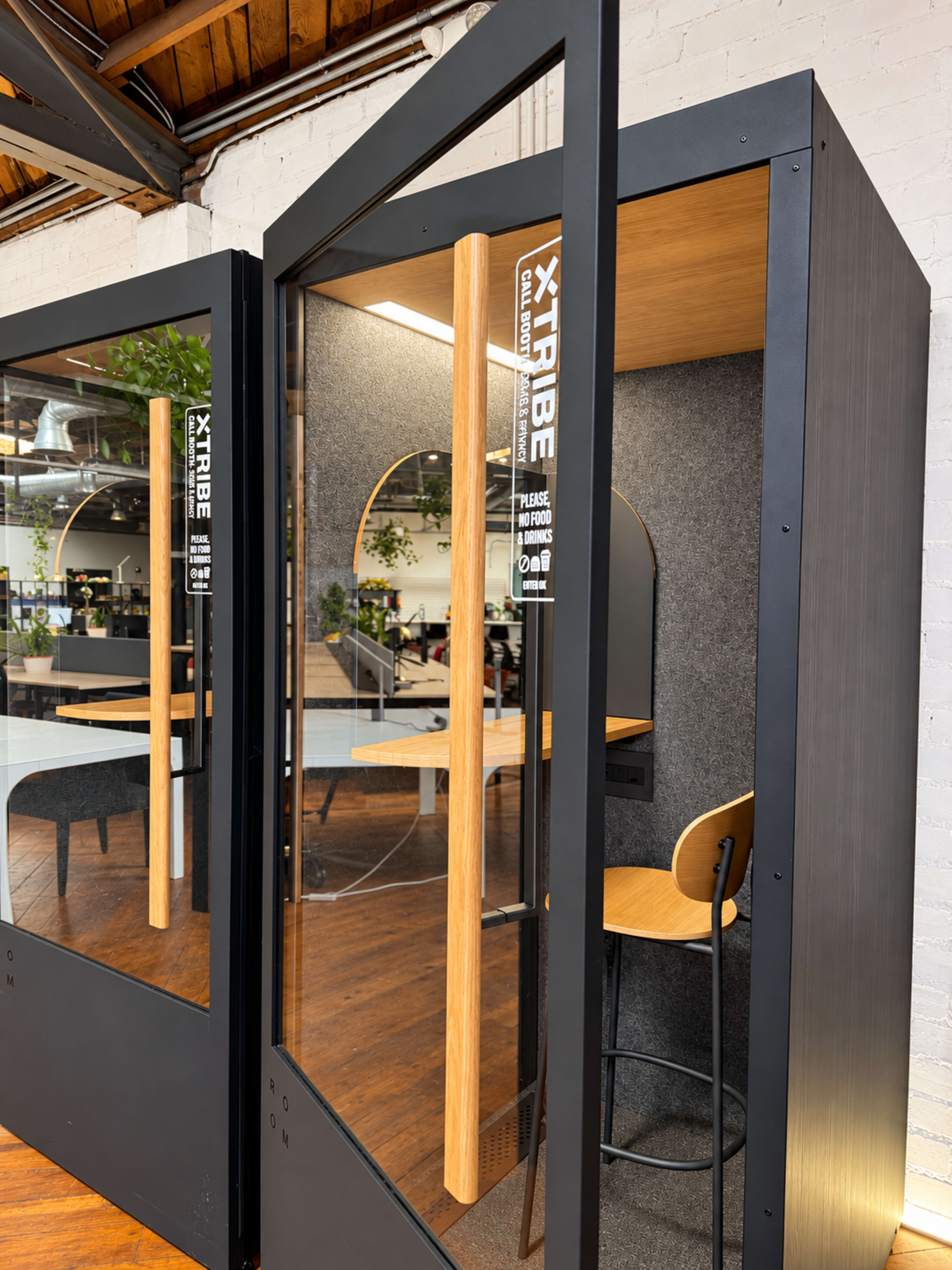 The image shows modern soundproof phone booths with glass doors, wooden accents, and a small table and chair inside, set in a contemporary office space.