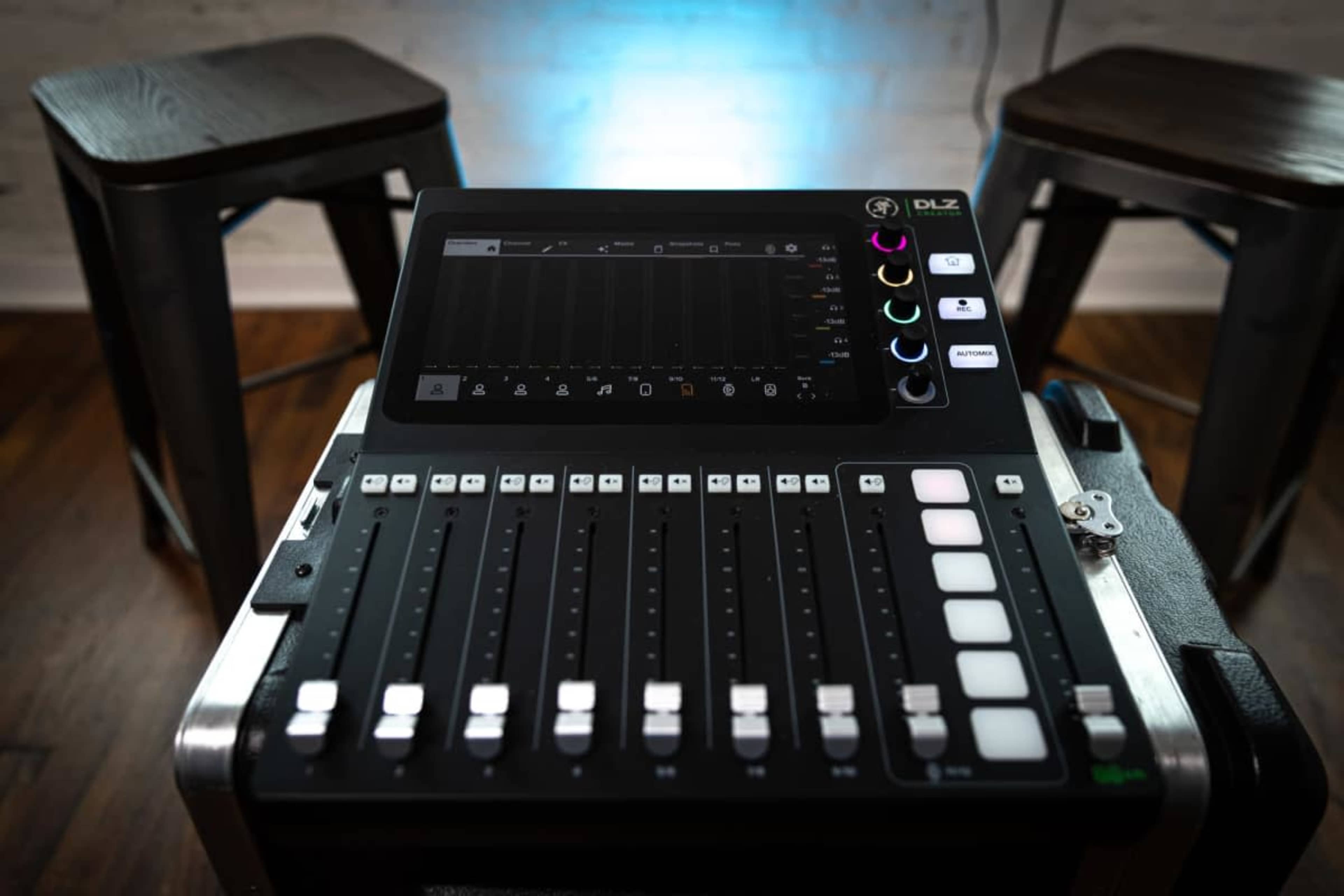 The image shows a digital mixing console with multiple sliders and buttons, placed on a black case in front of two wooden bar stools.