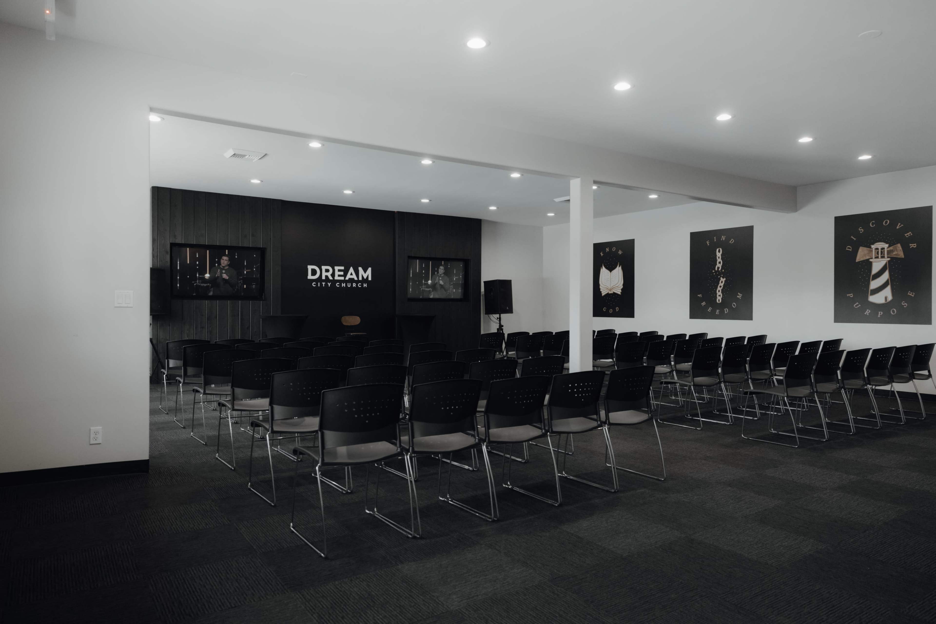 A conference room with rows of black chairs facing a stage that features a large sign reading "DREAM" and two artworks on the walls.