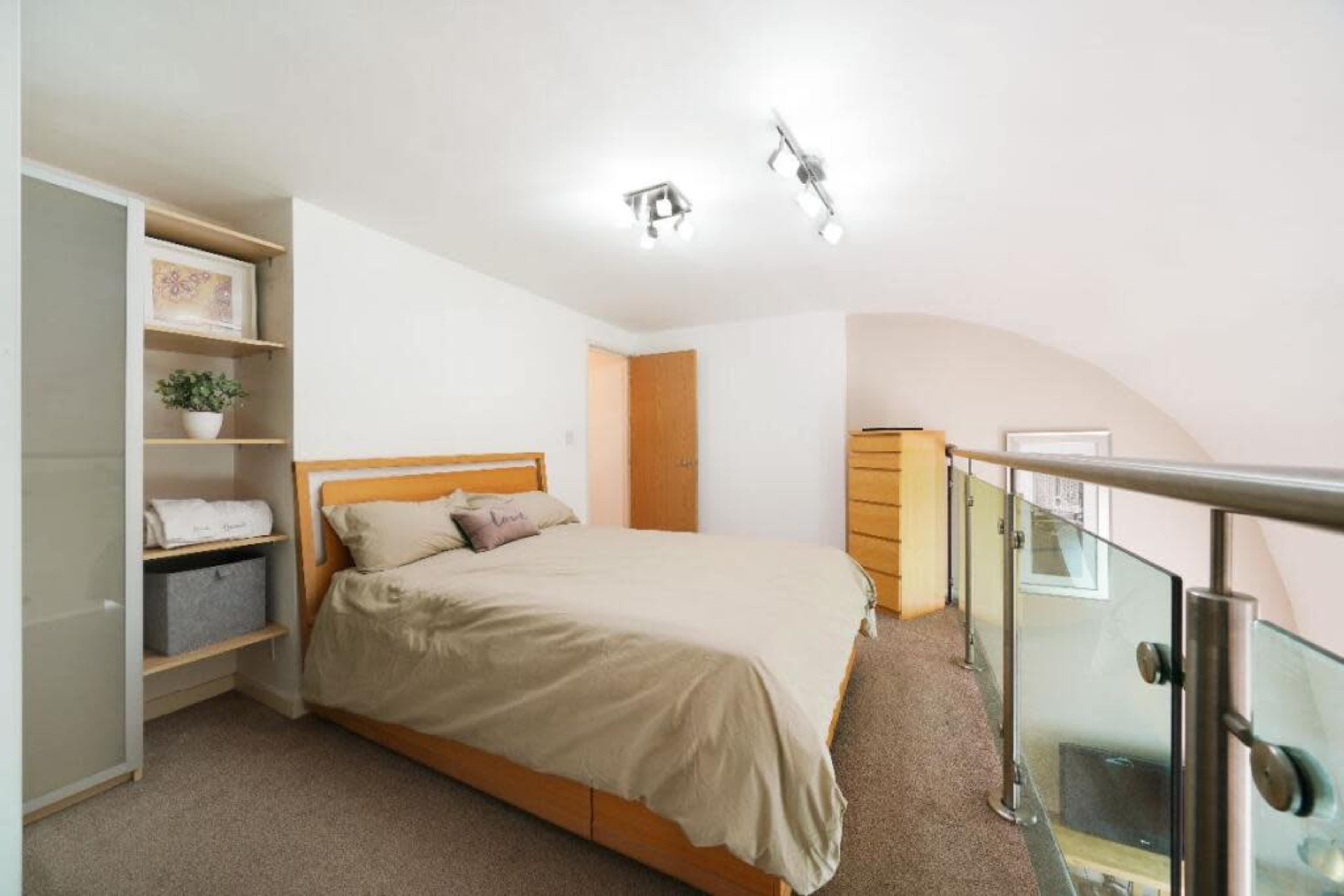 The image shows a simple bedroom featuring a wooden bed with beige bedding, a built-in wardrobe, and a small shelf with a plant.
