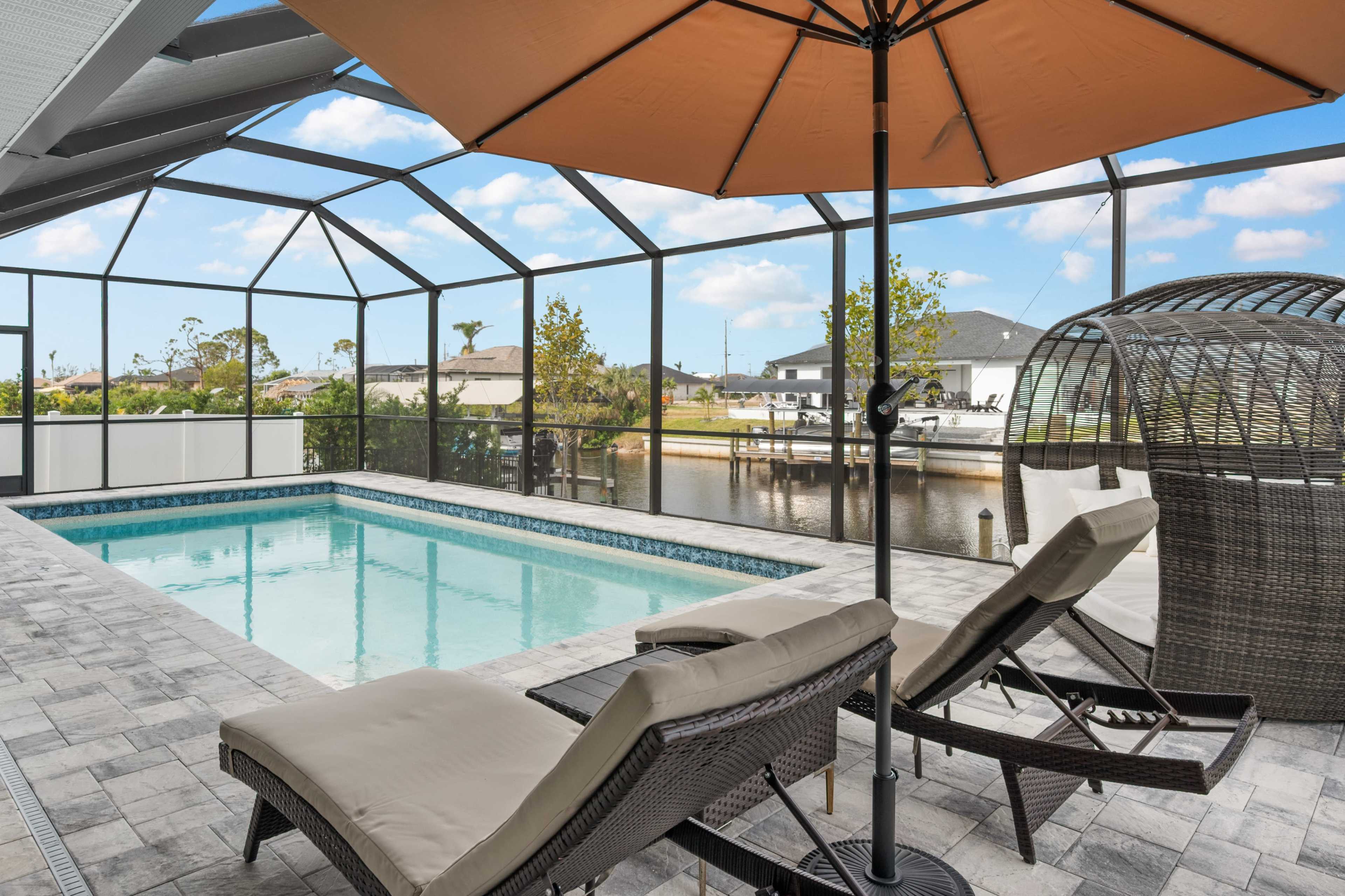 A screened-in patio area featuring a swimming pool, two lounge chairs, and an outdoor seating structure overlooking a canal.