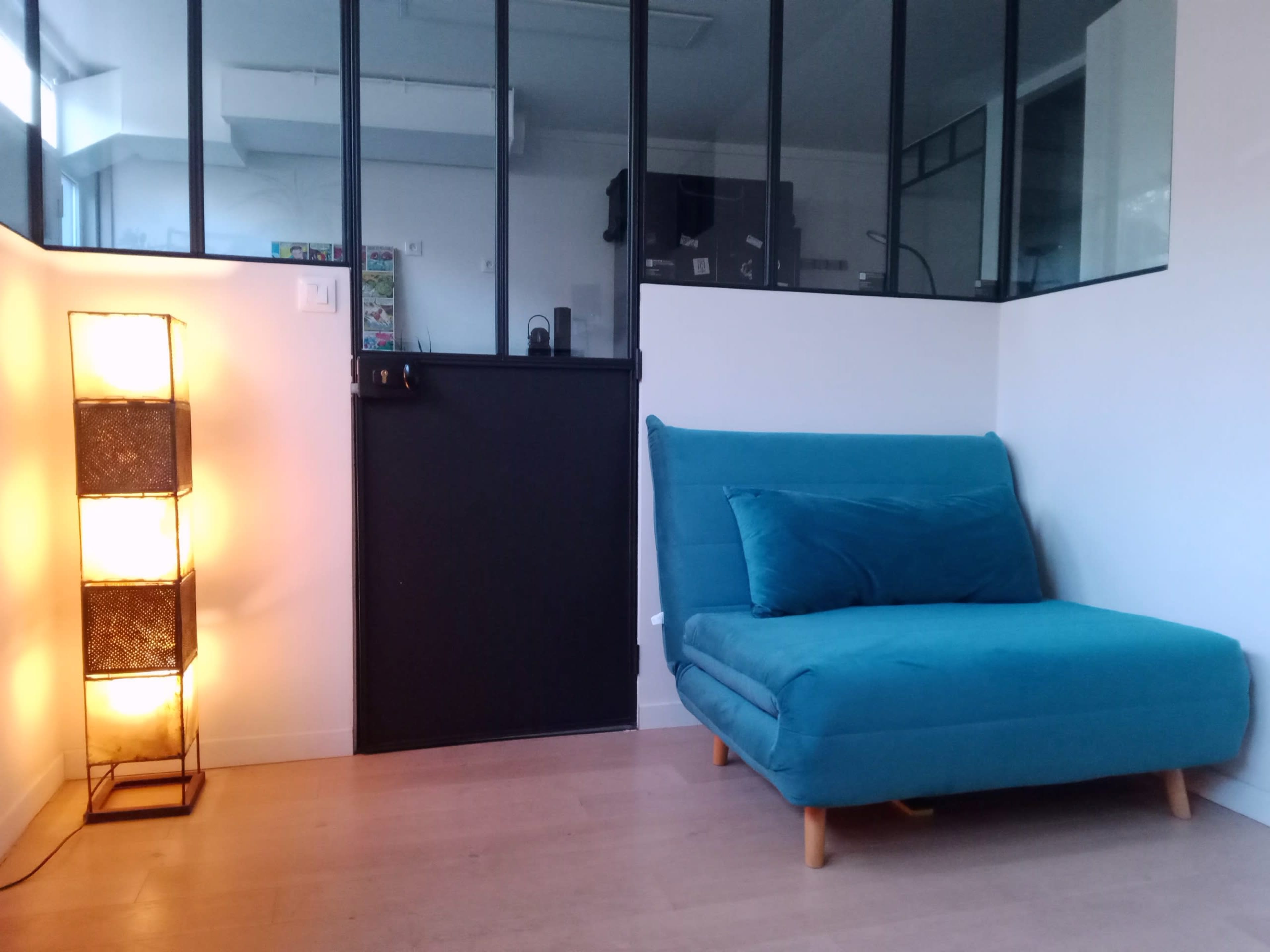 The image shows a modern interior space with a teal sofa and a tall, illuminated floor lamp next to a black door in a room with large windows.