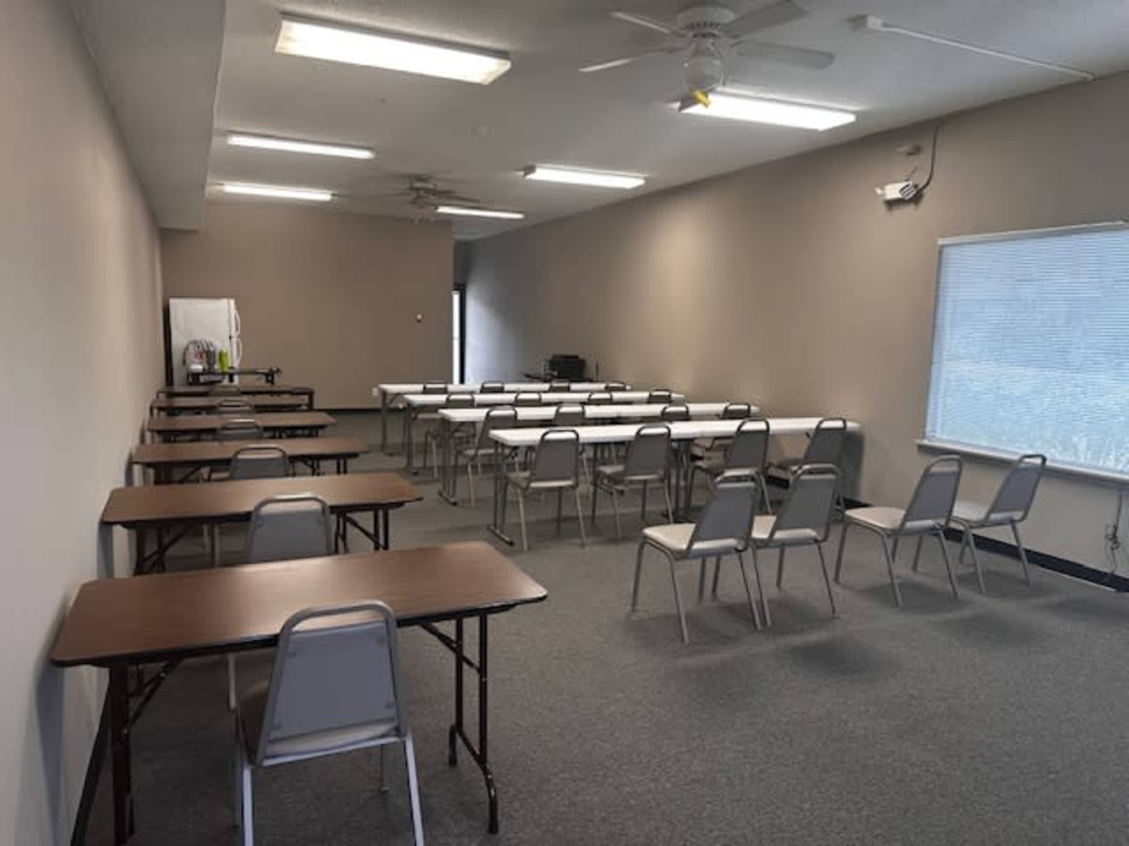 A spacious classroom is set up with multiple tables and chairs arranged in rows, and there is a window letting in natural light.