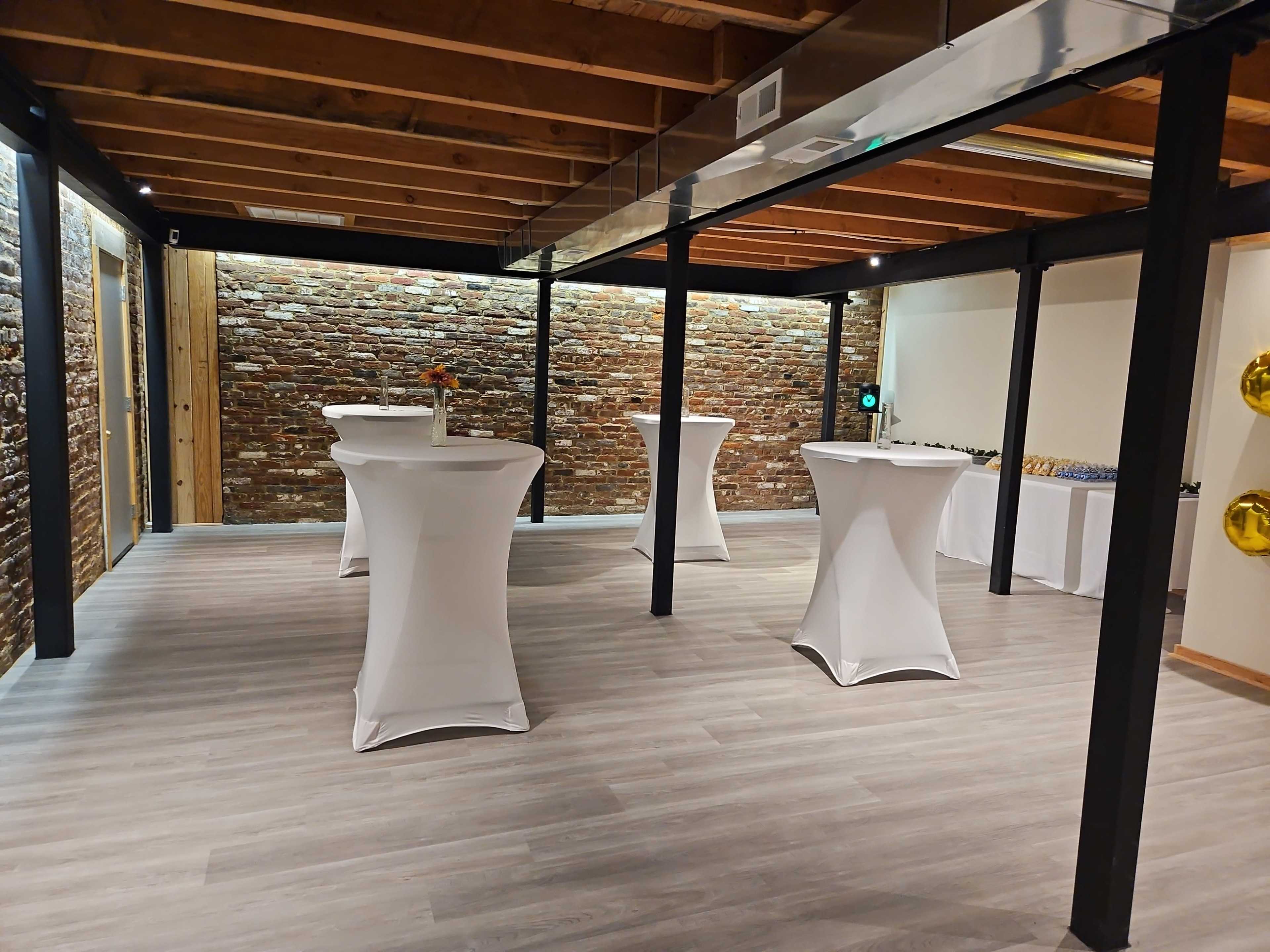 A spacious event room with exposed brick walls, wooden beams, and several cocktail tables draped in white linen.