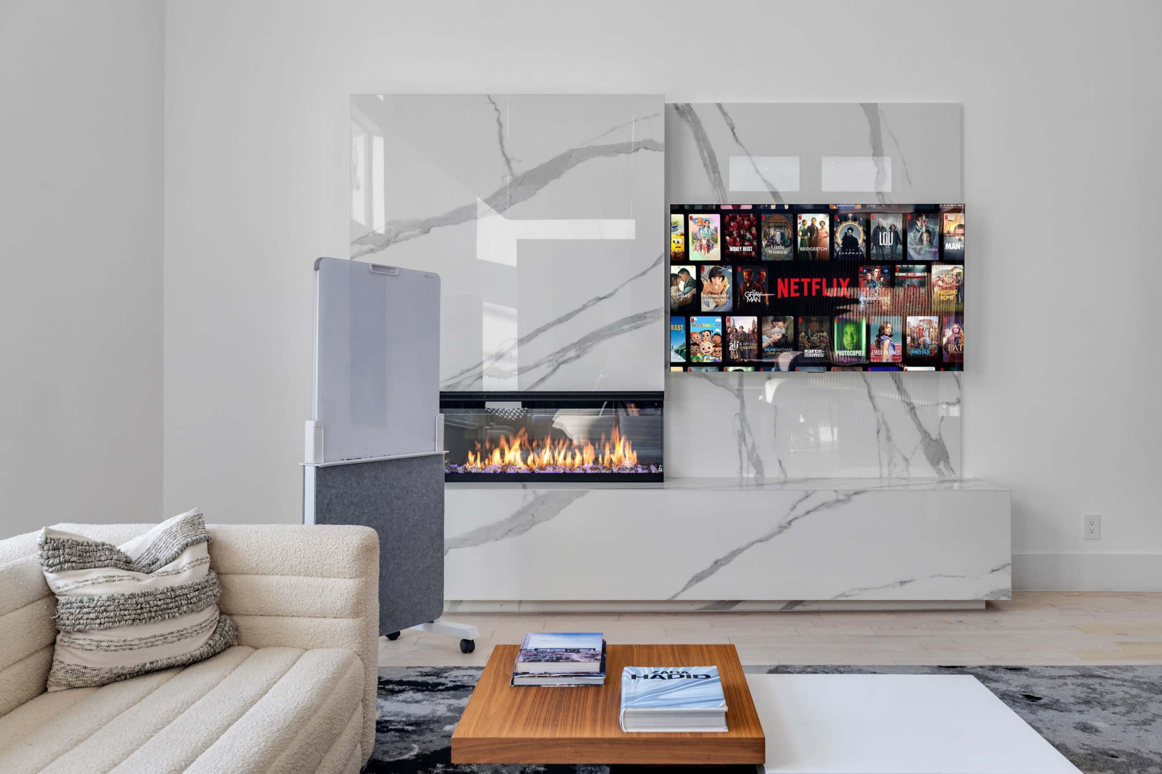The scene features a modern living room with a sleek marble accent wall, a built-in fireplace, and a television displaying a Netflix menu, accompanied by a light-colored sofa and a wooden coffee table.