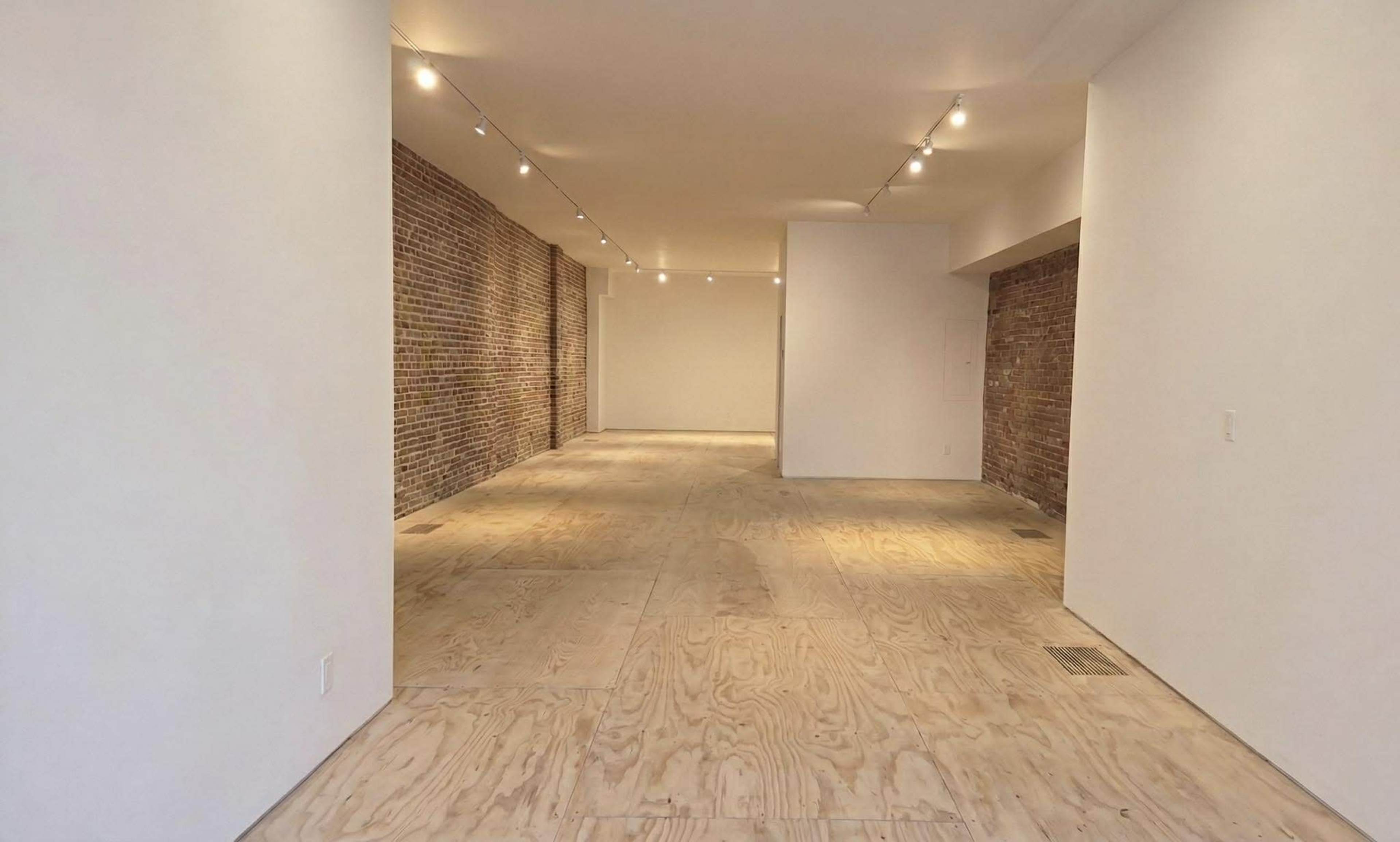 The image shows a spacious, unfinished interior with exposed brick walls and a plywood floor, illuminated by track lighting.
