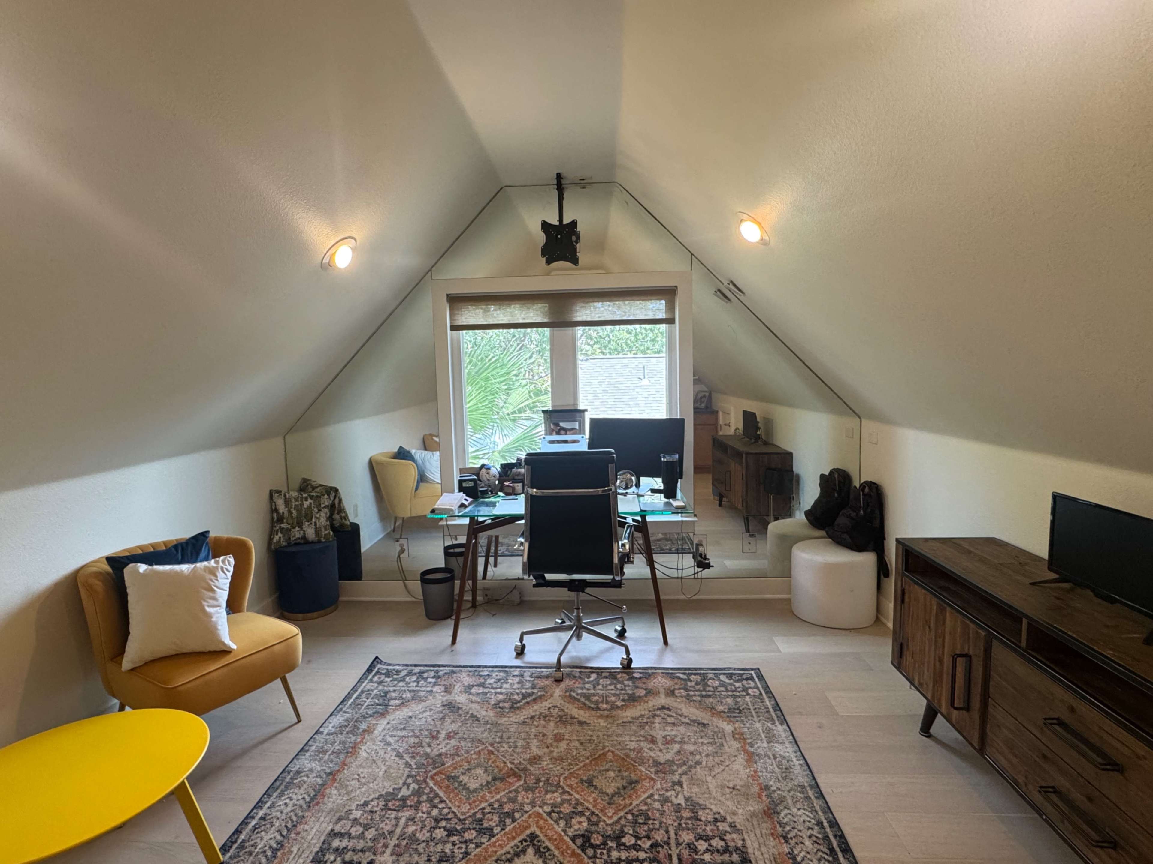 A spacious attic office features a desk with a computer, a comfortable chair, and a rug, alongside various furnishings and a large window.