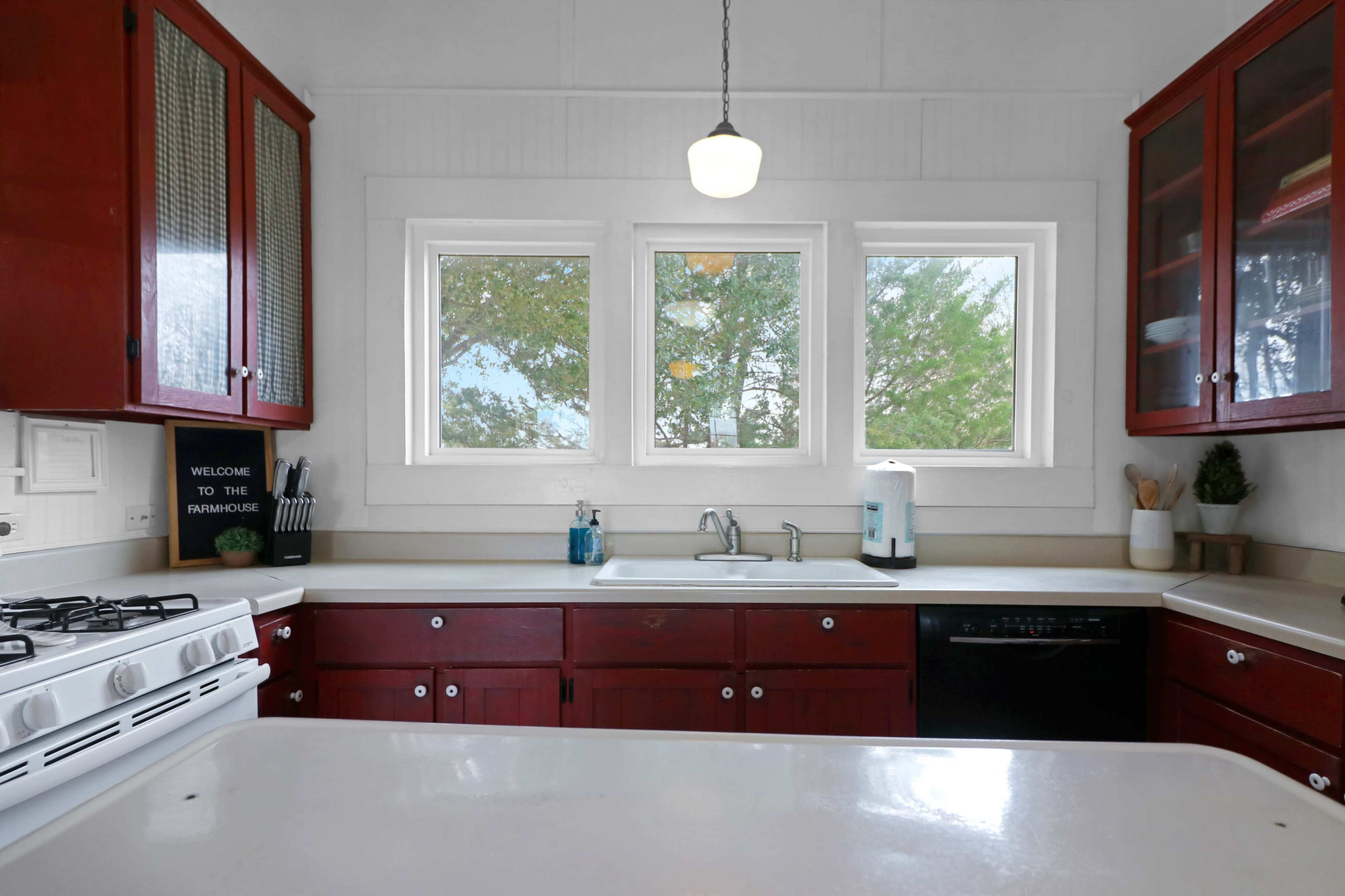 A kitchen features red cabinets and a white countertop, with a view through three windows overlooking greenery.