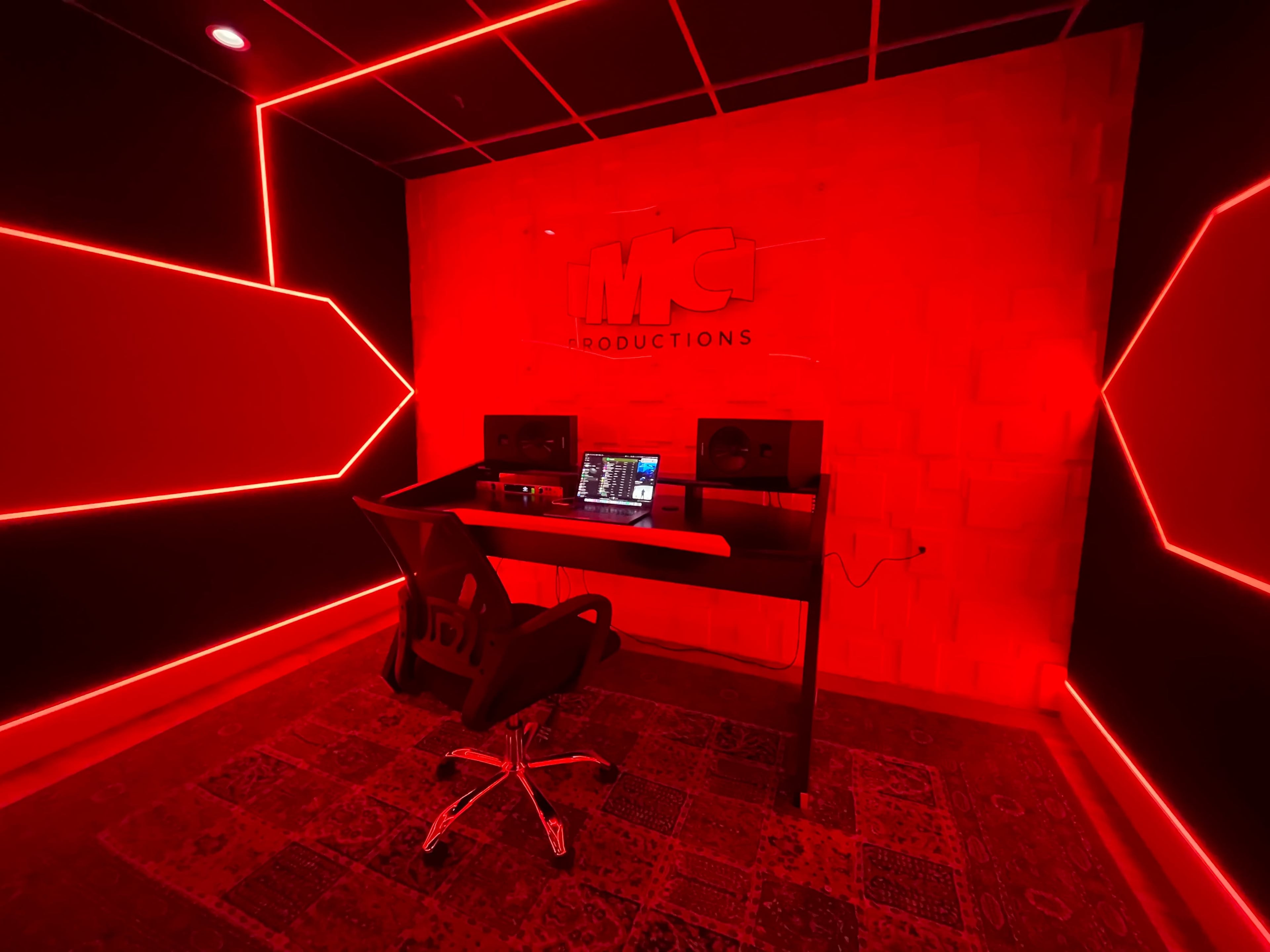 A music production studio features a black wall with red LED lighting, a desk with a laptop, and two studio monitors.