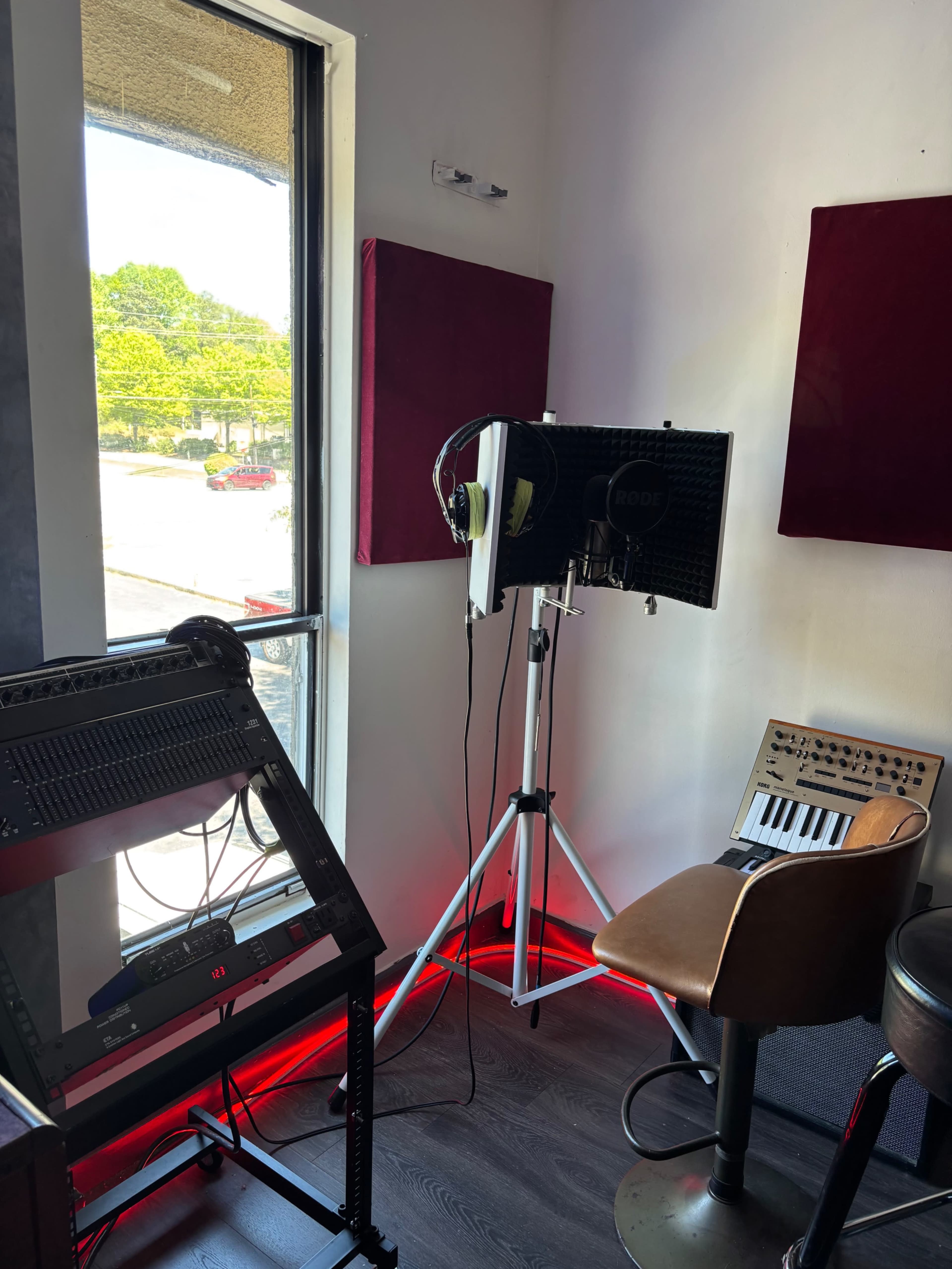 The image shows a small music recording space with soundproofing panels on the wall, a microphone on a stand, and a keyboard setup by a window.