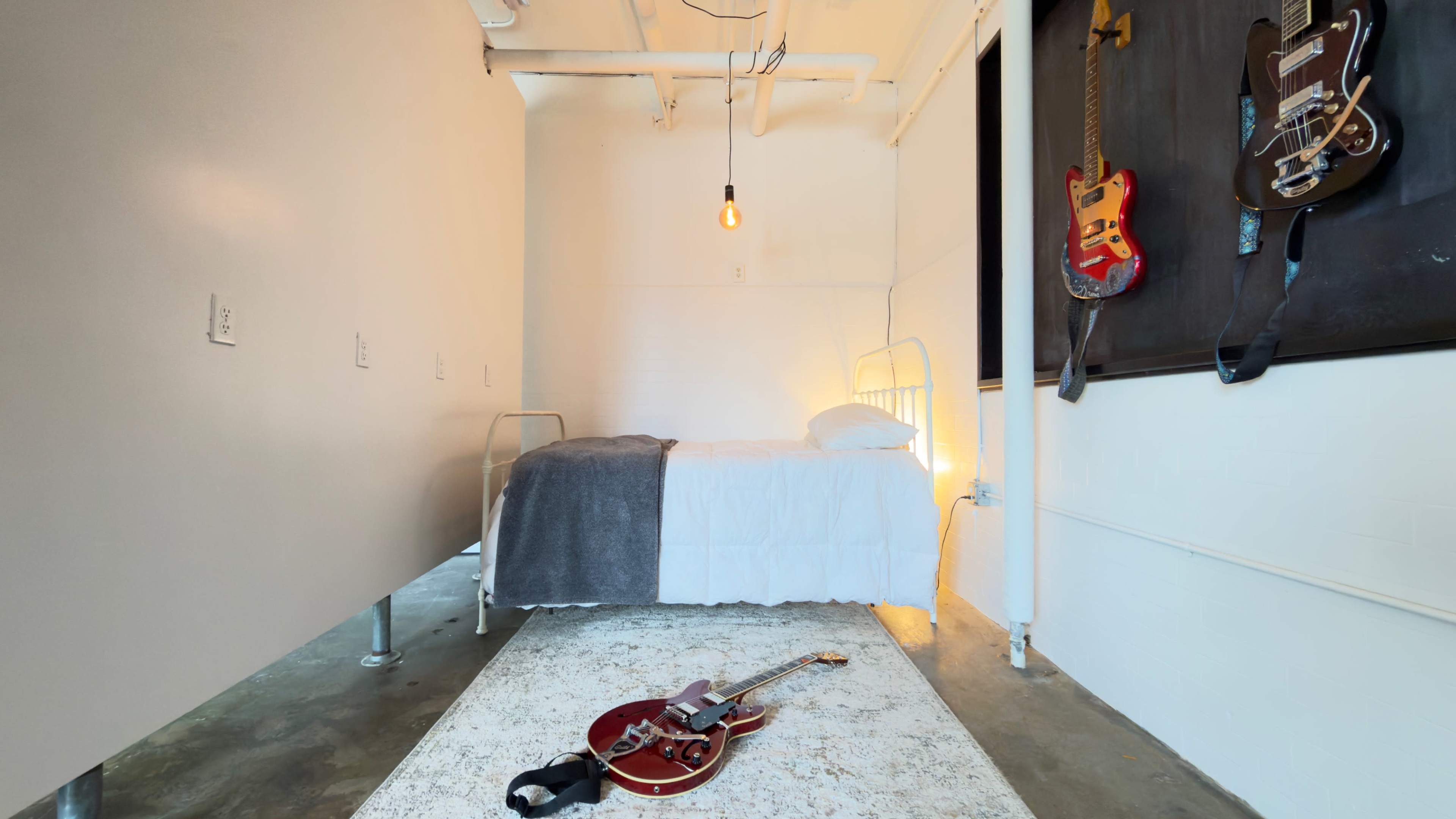 The image shows a minimalist bedroom with a single bed, a concrete floor, and an electric guitar lying on a rug.
