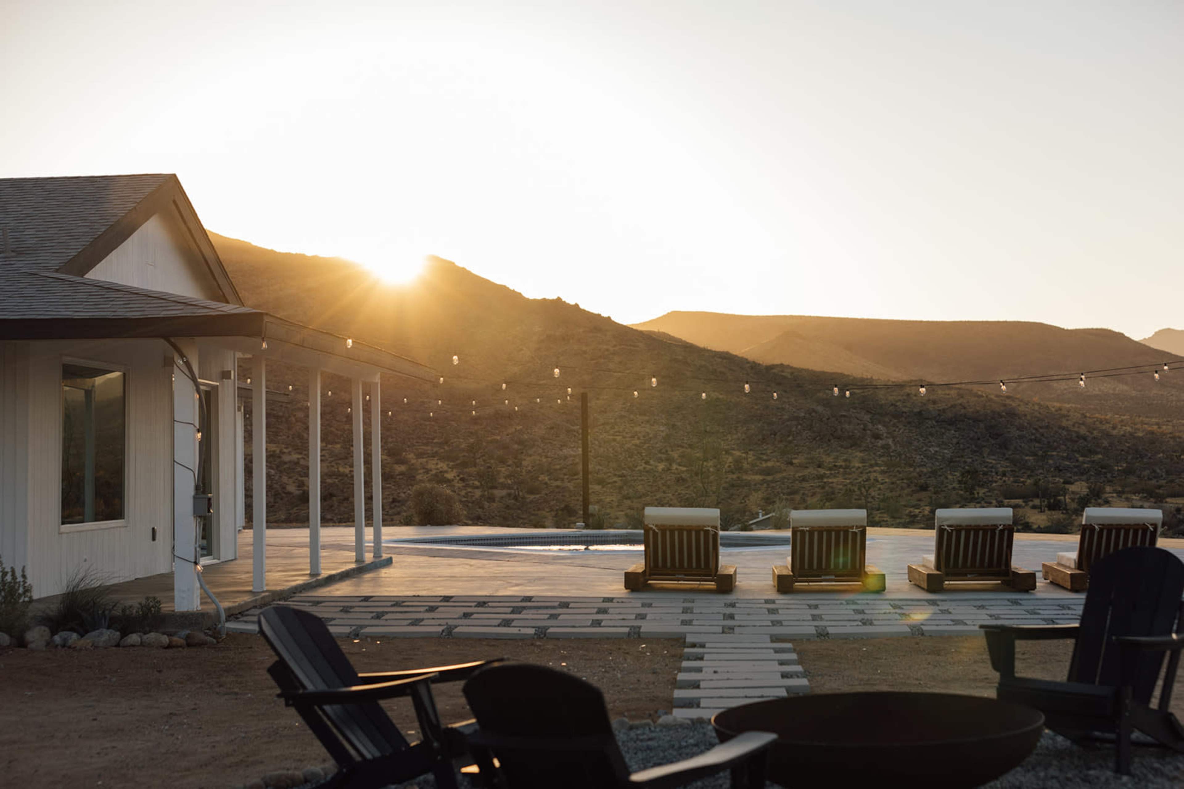 A modern house is situated in a rocky landscape, with wooden lounge chairs arranged near a patio under a setting sun.
