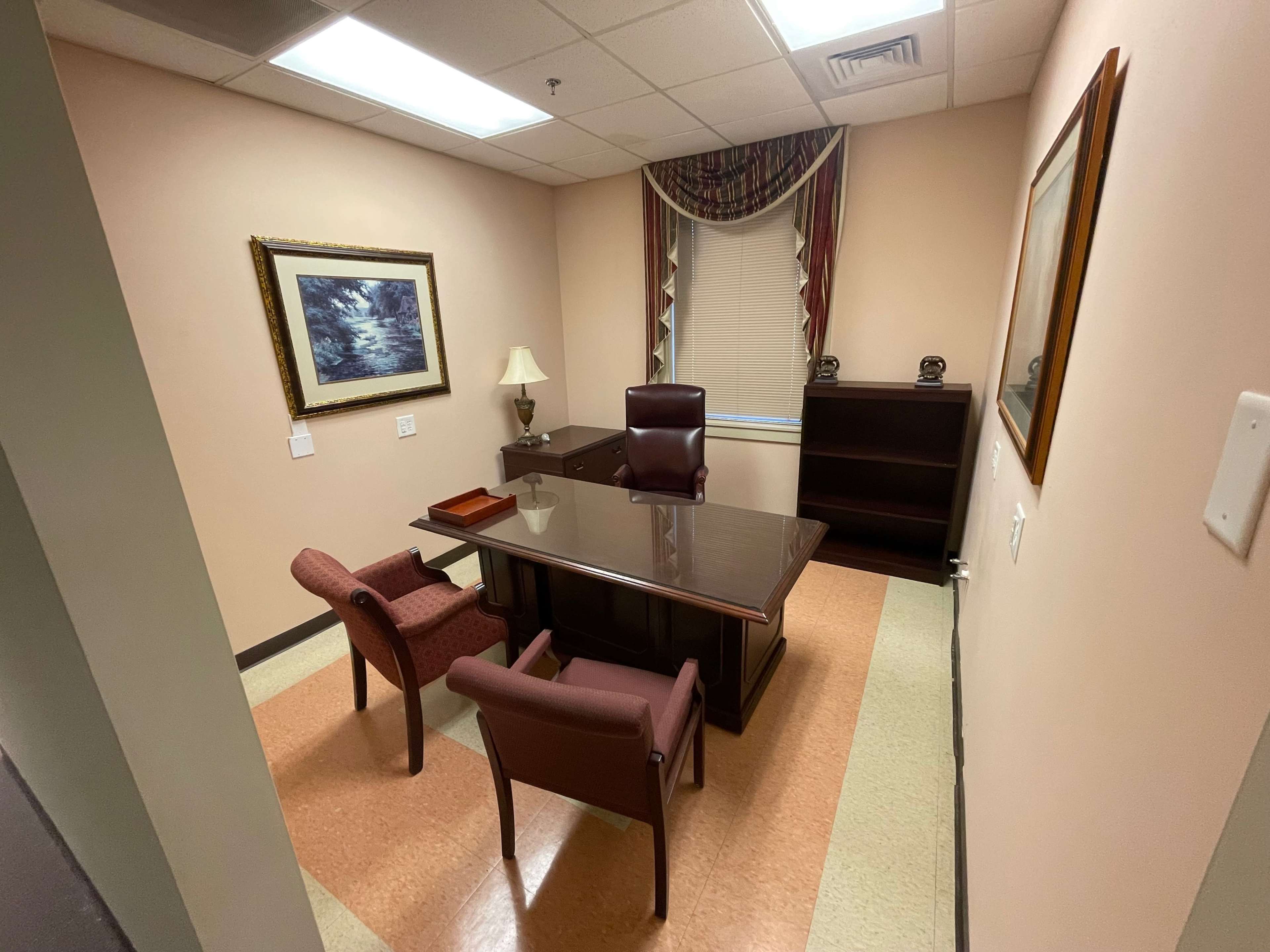 A small office featuring a wooden desk, two chairs, a lamp, and framed artwork on the walls.
