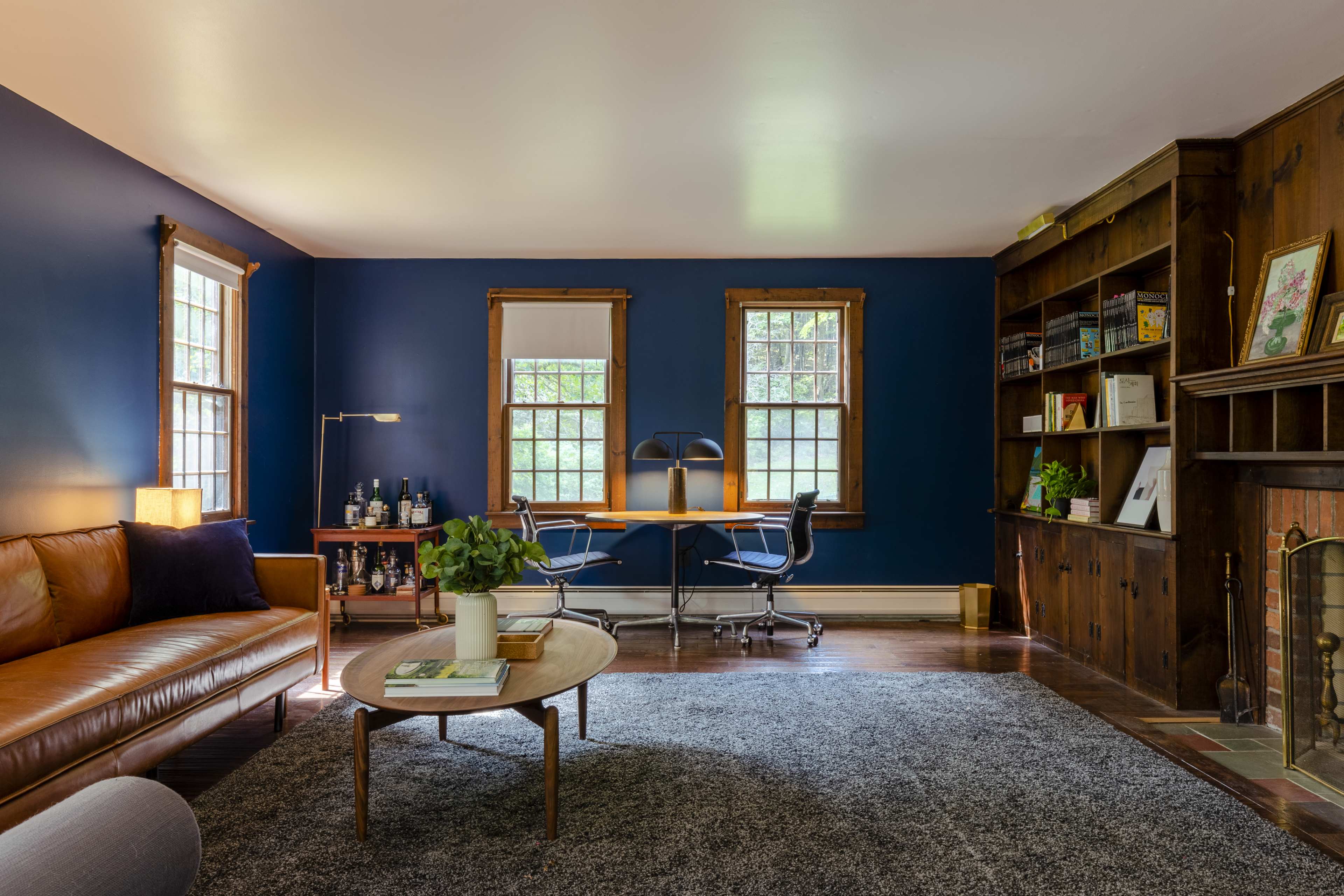 A cozy living room features a brown leather sofa, a round coffee table, and a desk by two windows, with a bookshelf and a fireplace in the background.