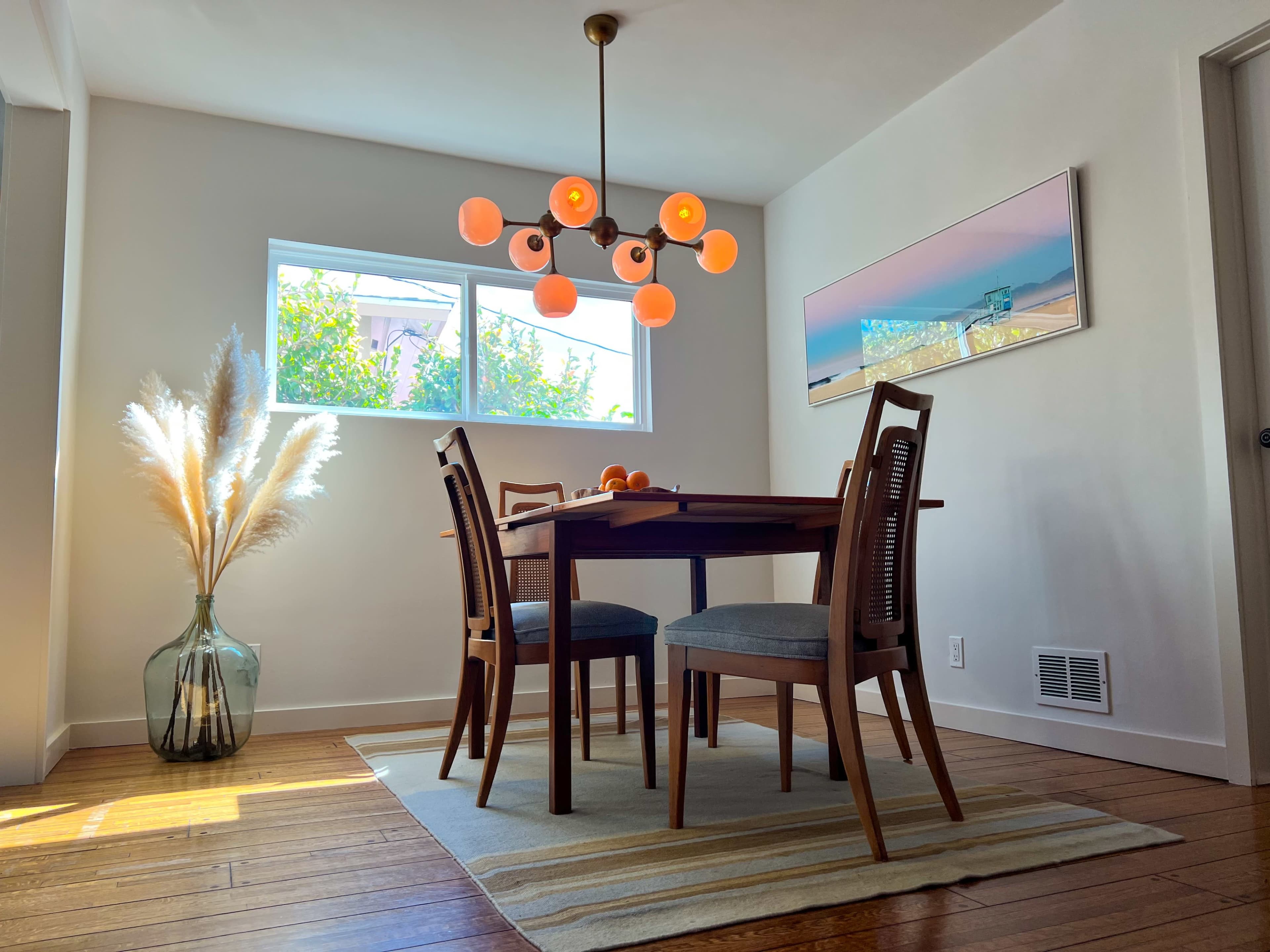 A dining area features a table with four chairs, a modern light fixture, a large plant in a glass vase, and two windows allowing natural light.